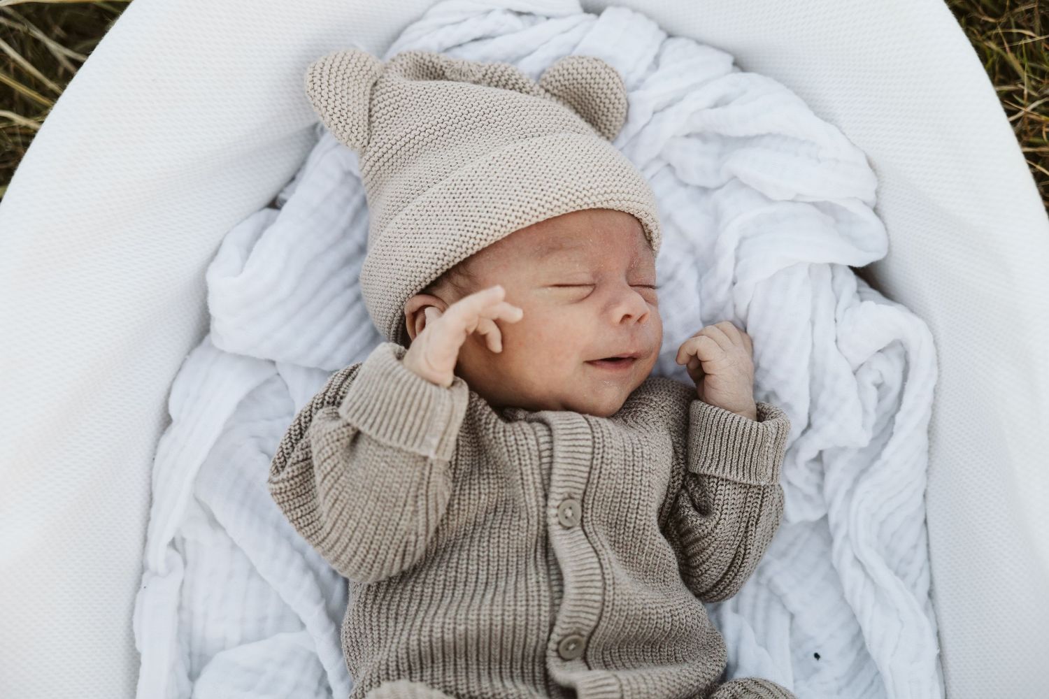Peaceful newborn baby sleeping in white bassinet wearing a beige knit hat and romper.