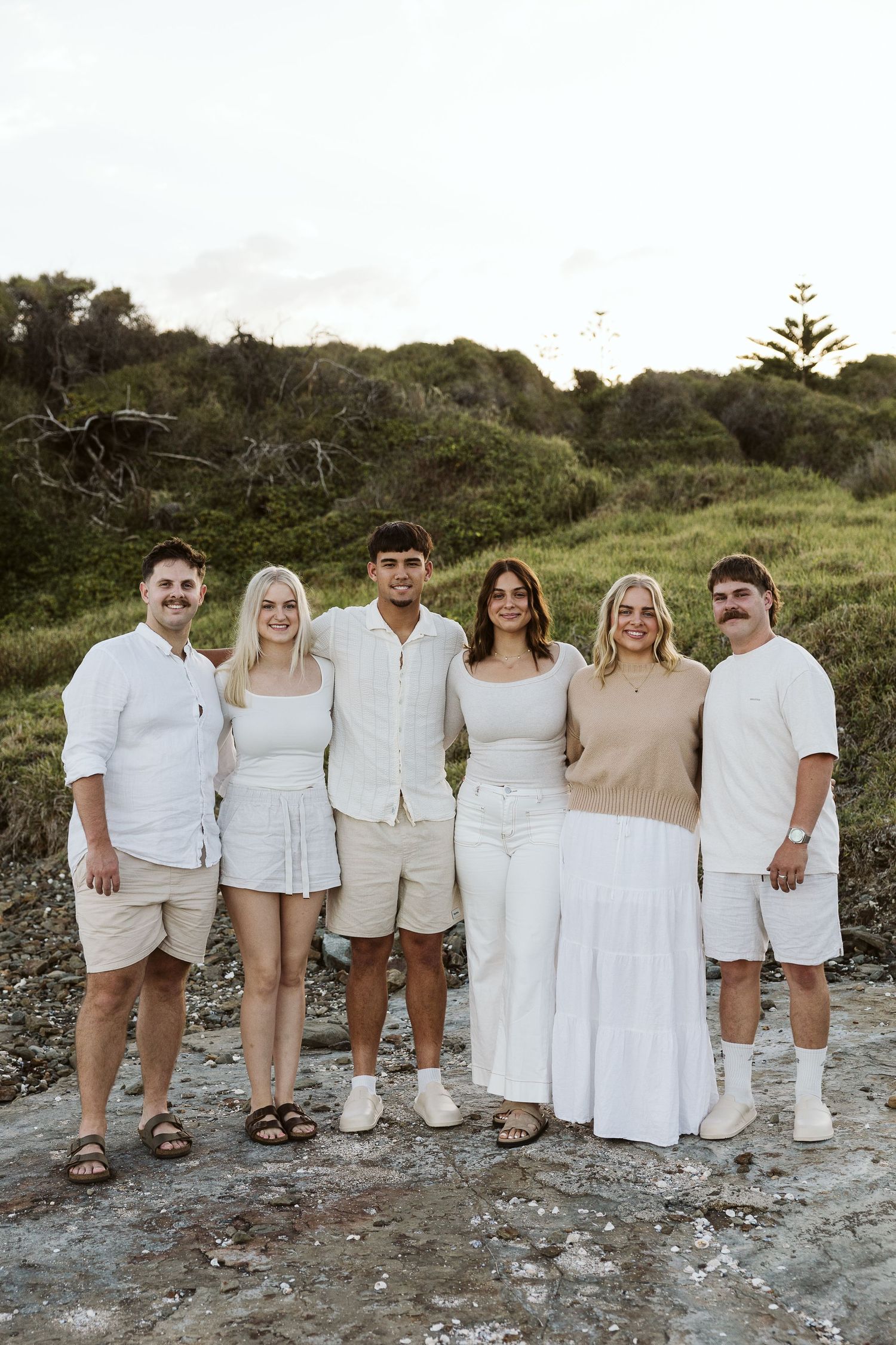 Six people in white outfits pose together on rocky coastal ground with lush green bushland backdrop.