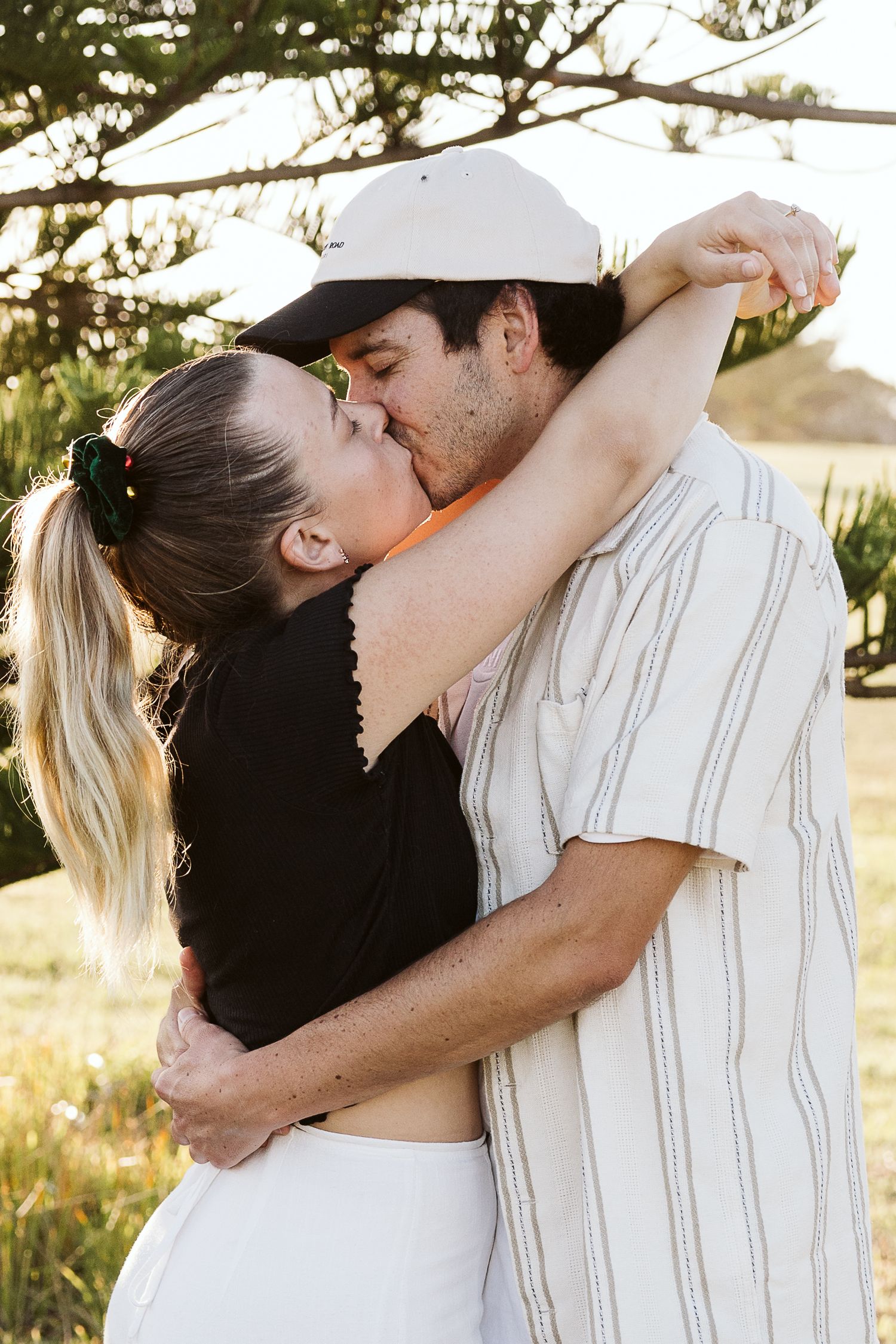 A couple shares a romantic kiss outdoors, embracing near a tree in warm golden sunlight.