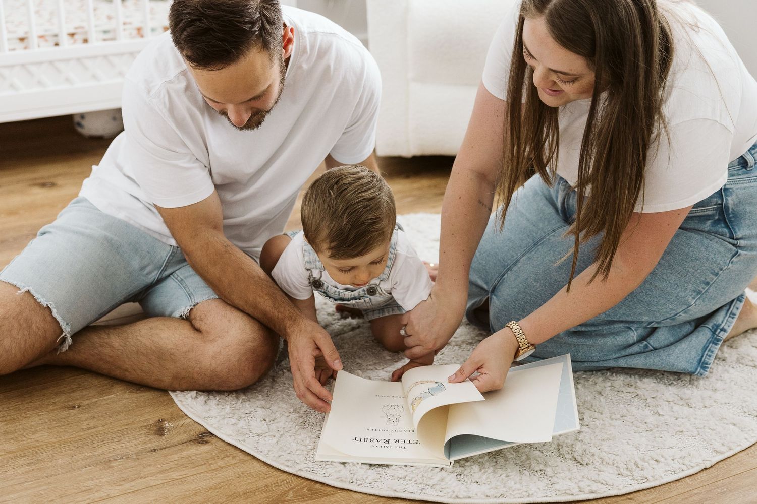 A young family sits on a rug reading a book together with their toddler on a hardwood floor.