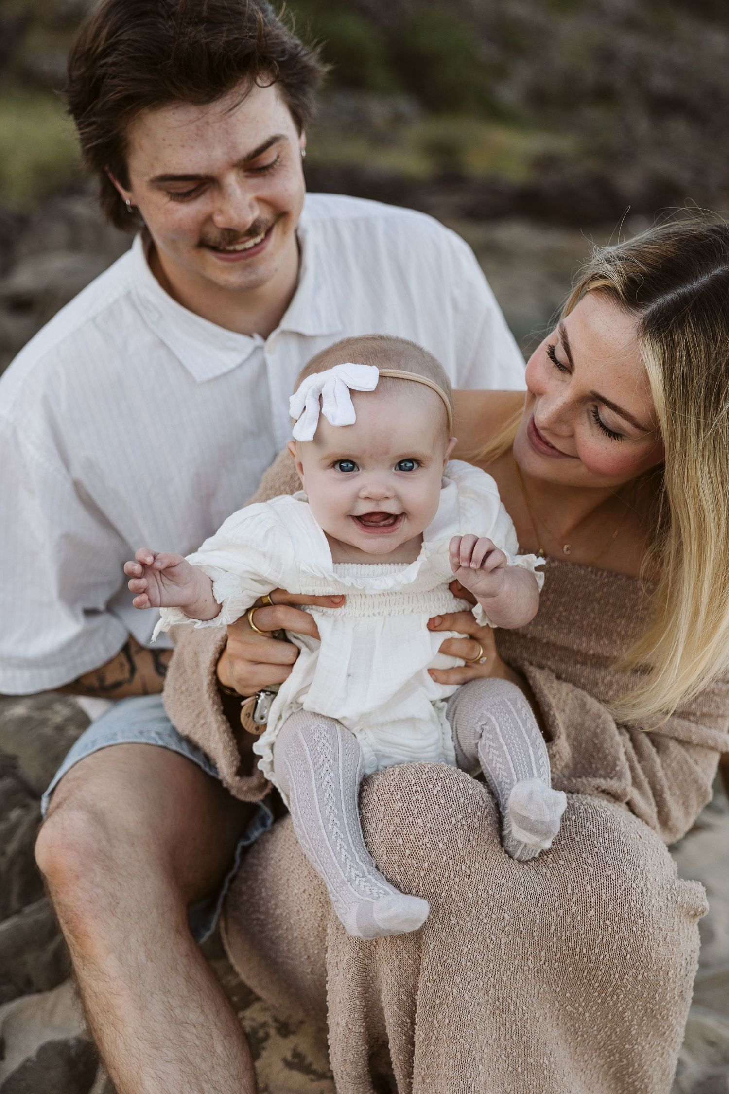 Happy young family with tattooed father and blonde mother holding smiling baby girl outdoors.