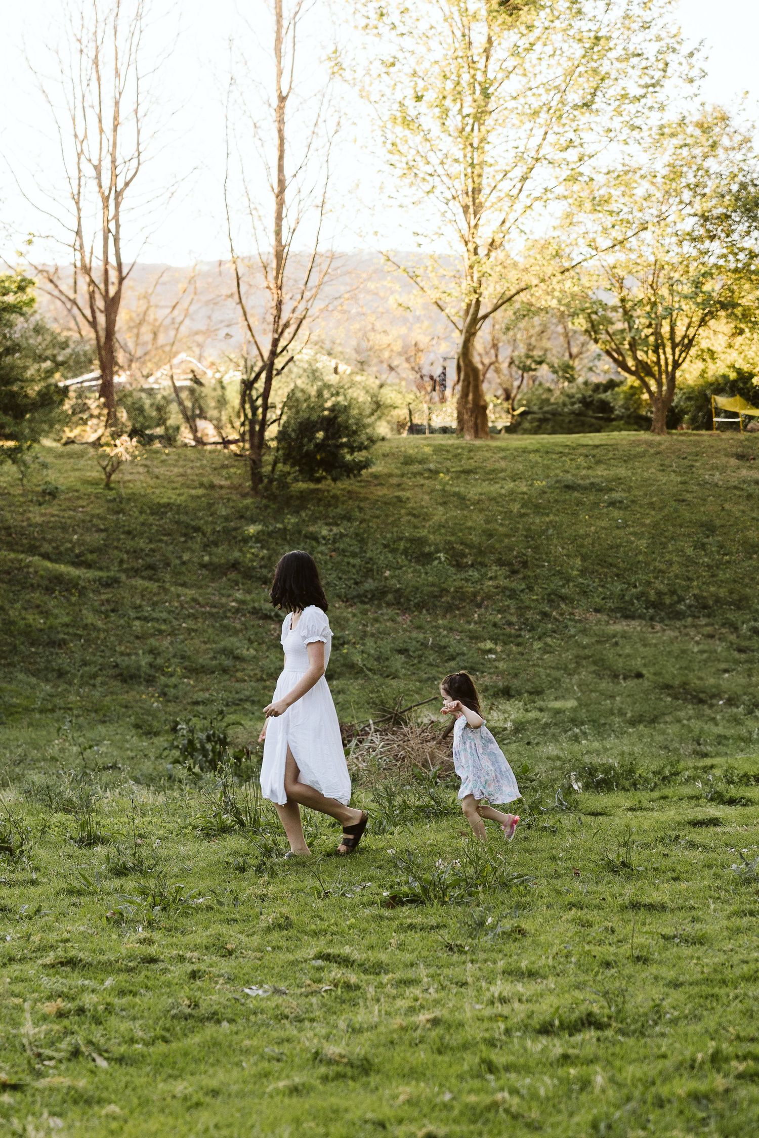 A mother and daughter in white dresses walk together through a lush green field surrounded by trees at golden hour.