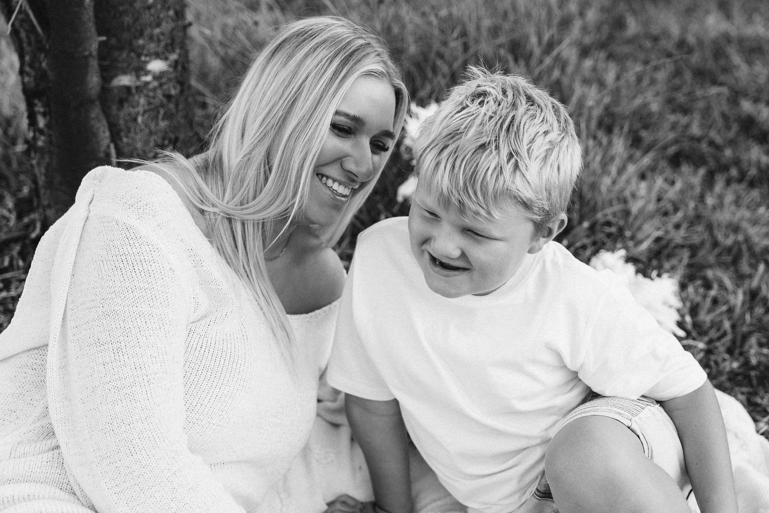 Blonde mother and young son laughing together while sitting outdoors in black and white portrait.