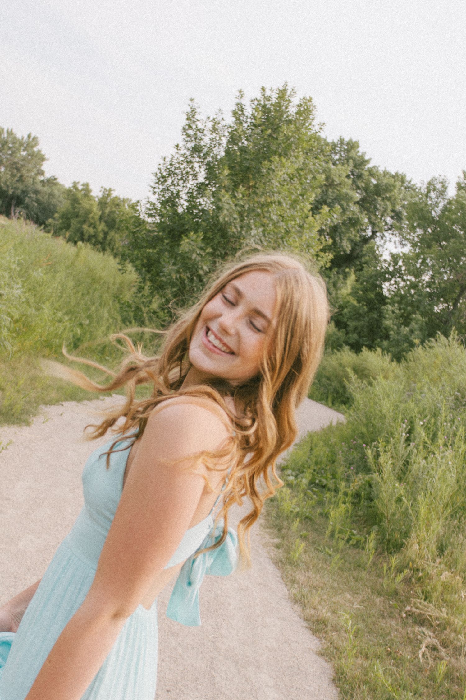 Young woman in mint dress smiling joyfully on a nature path surrounded by lush green trees.