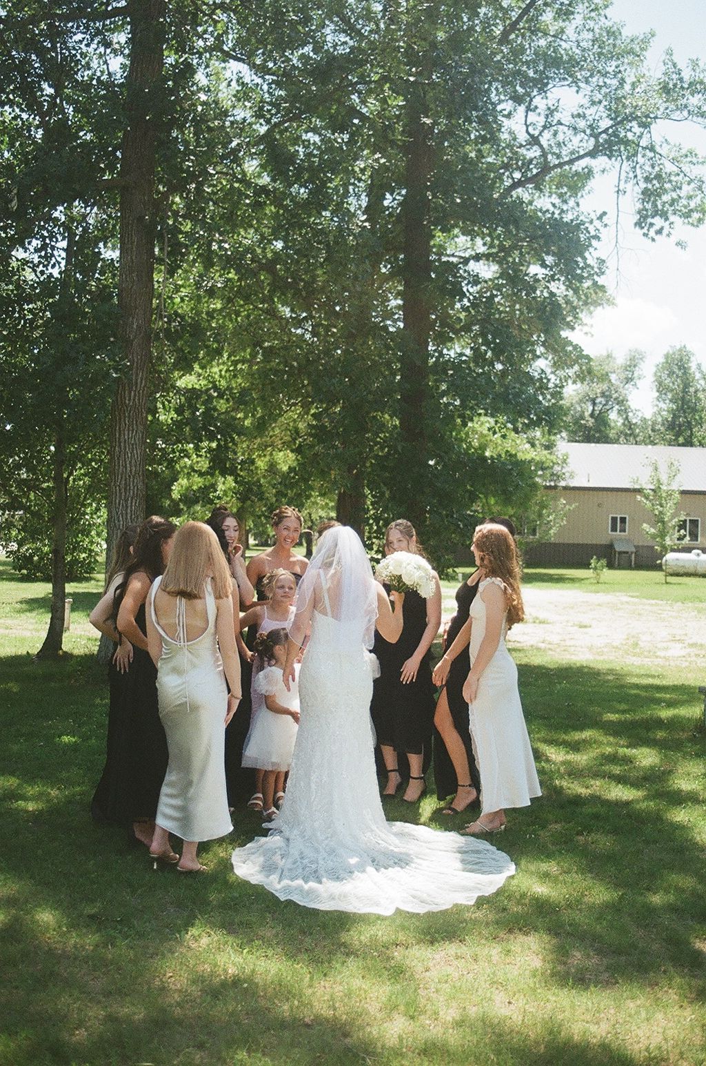 Bride in white gown with veil surrounded by bridesmaids in black and white dresses under tall trees.