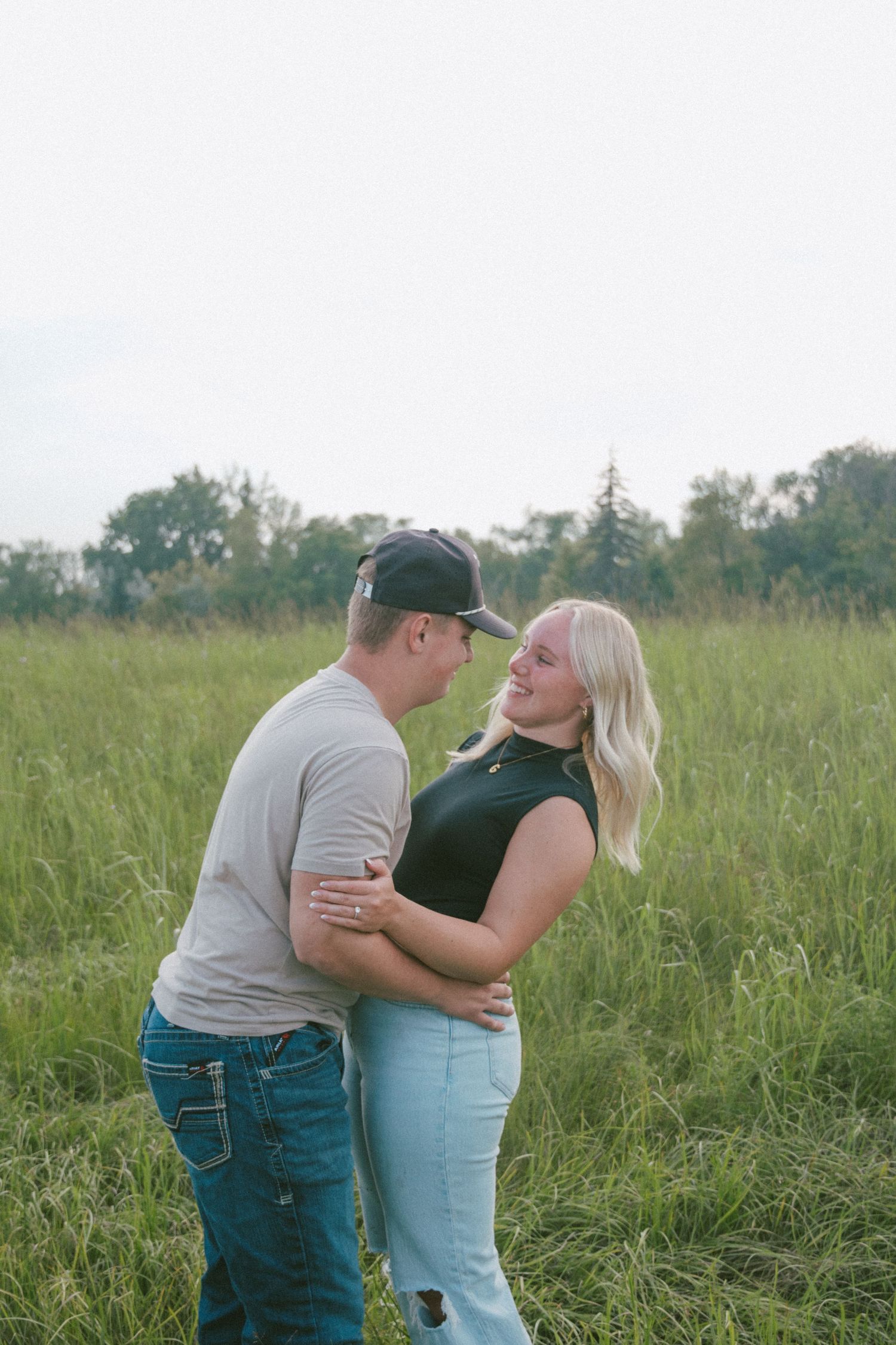 Couple embracing and smiling at each other in a tall green grassy field at dusk.