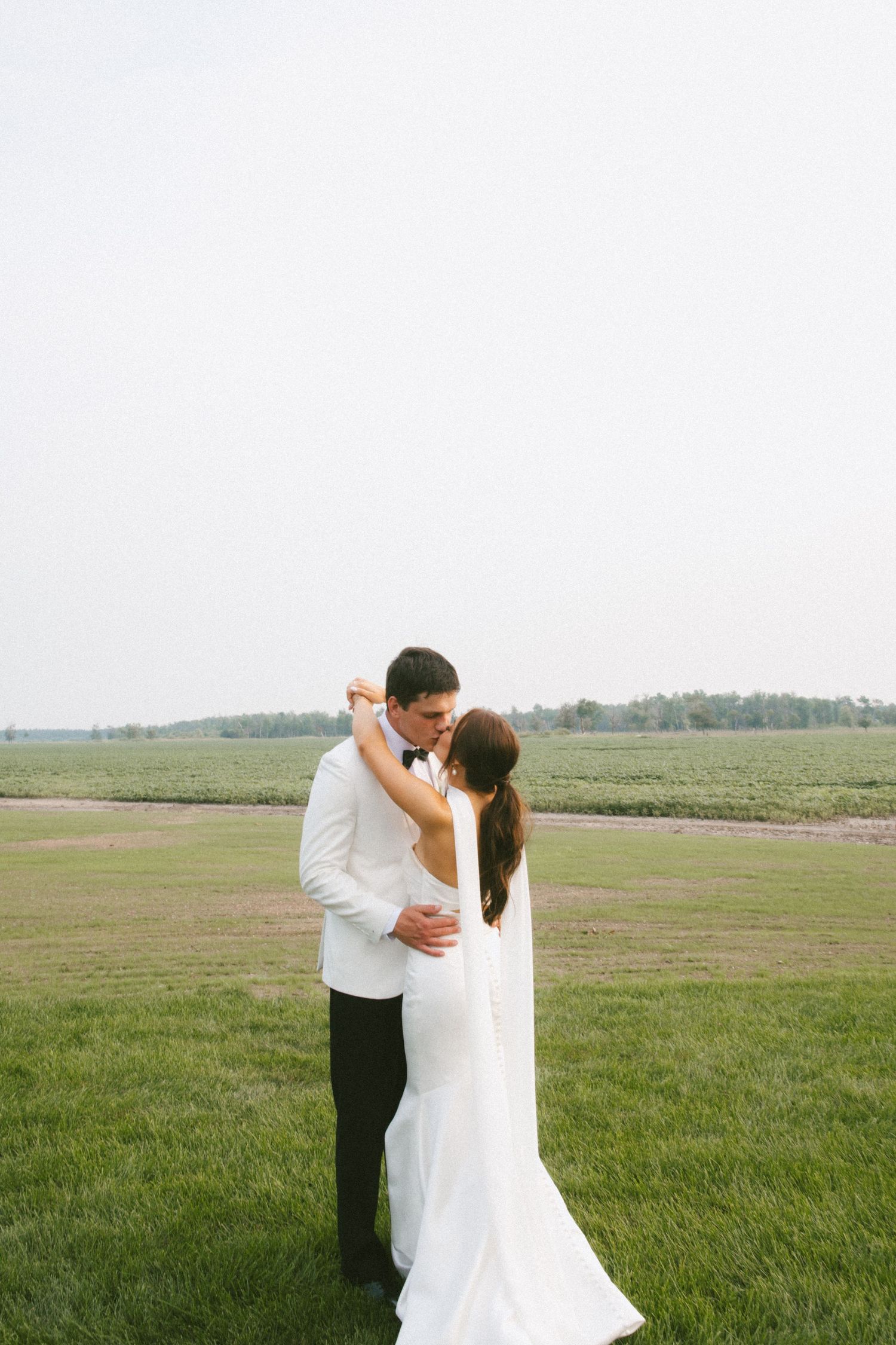Bride and groom share a tender kiss while dancing on a lush green field at sunset.