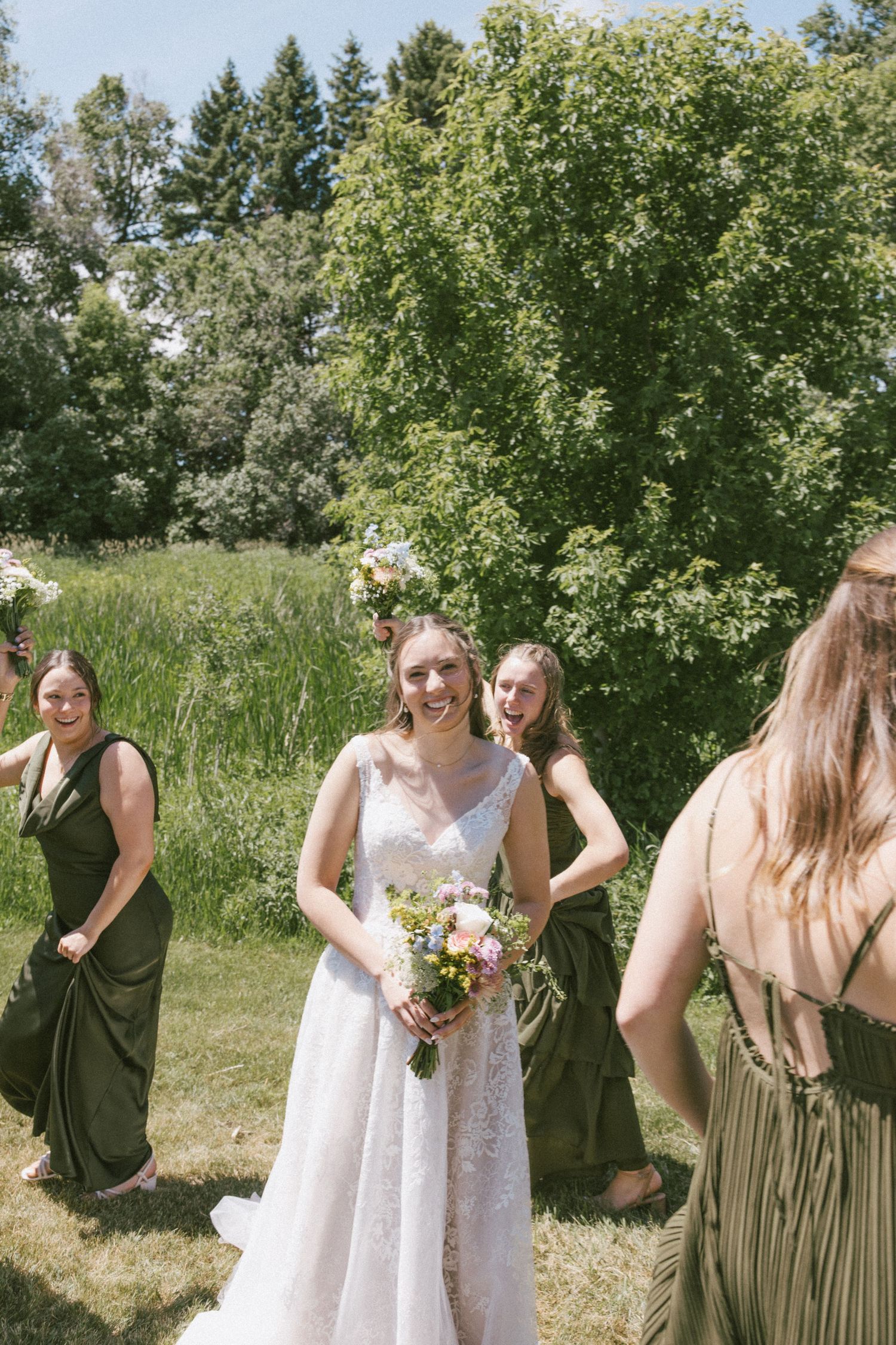 Bride in white lace gown holds floral bouquet surrounded by bridesmaids in olive green dresses outdoors.