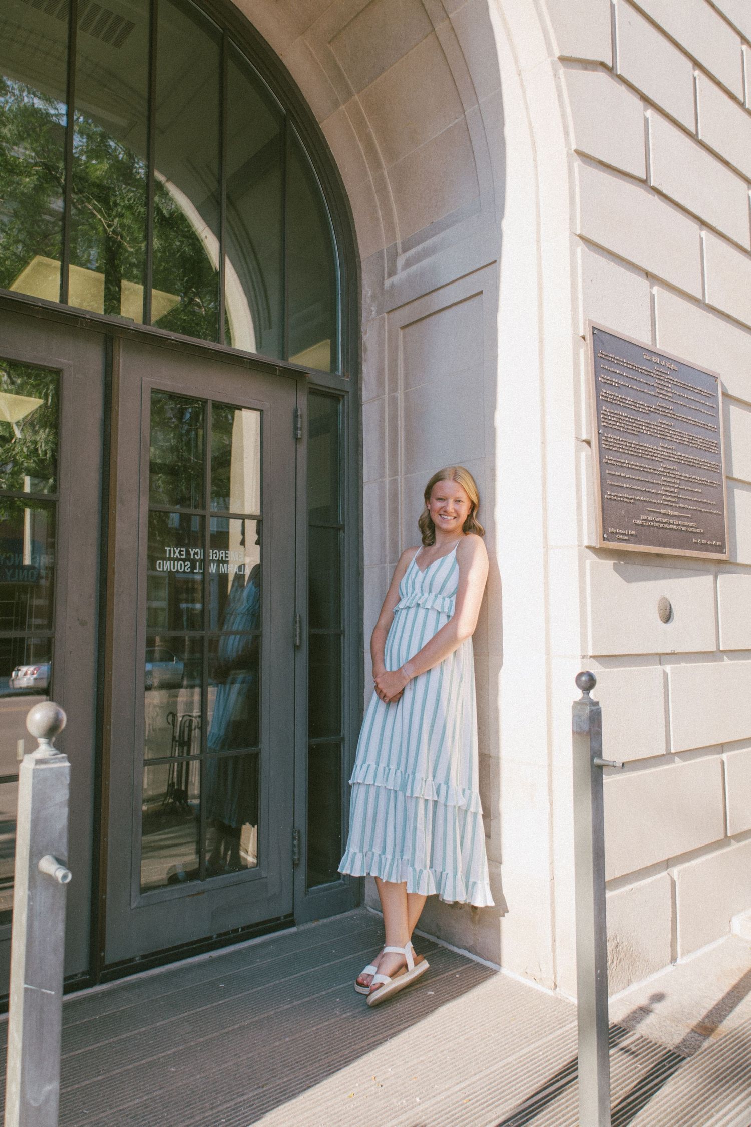 Smiling woman in striped midi dress posing by elegant arched doorway of historic stone building on sunny day.