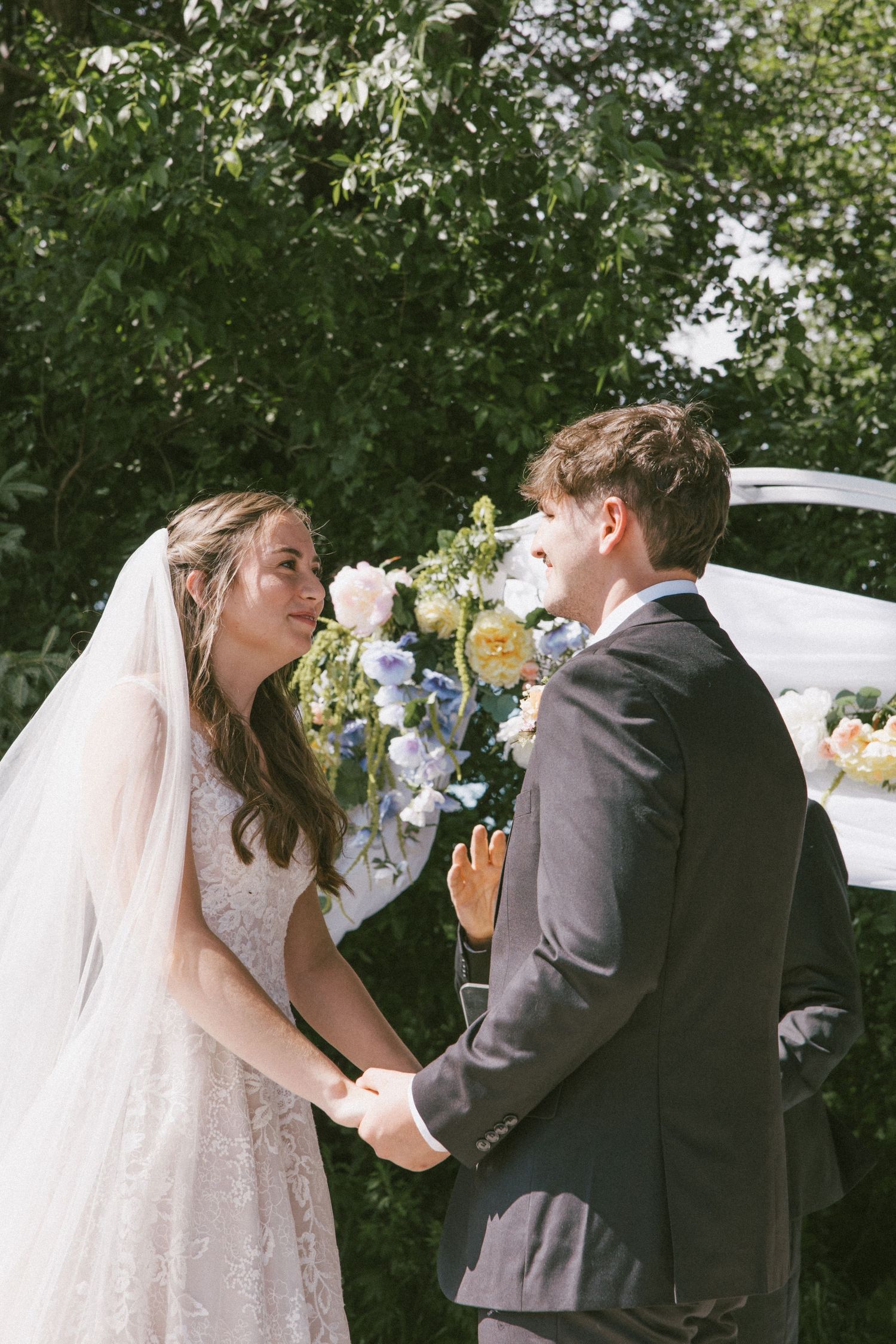 Bride and groom holding hands at outdoor wedding ceremony with floral arch and lush green trees in background.