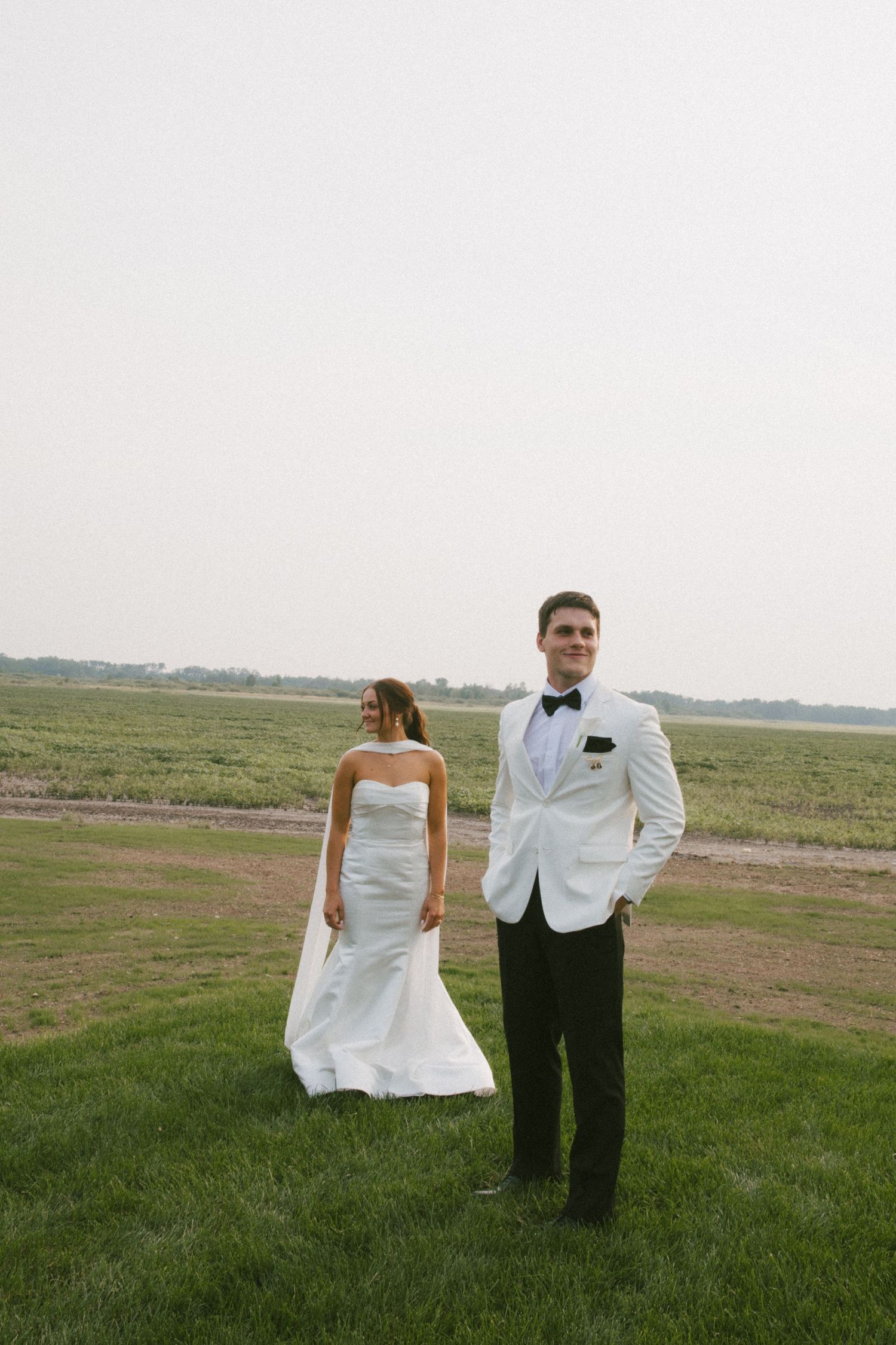 Bride in white strapless mermaid gown and groom in white tuxedo jacket pose on green field.