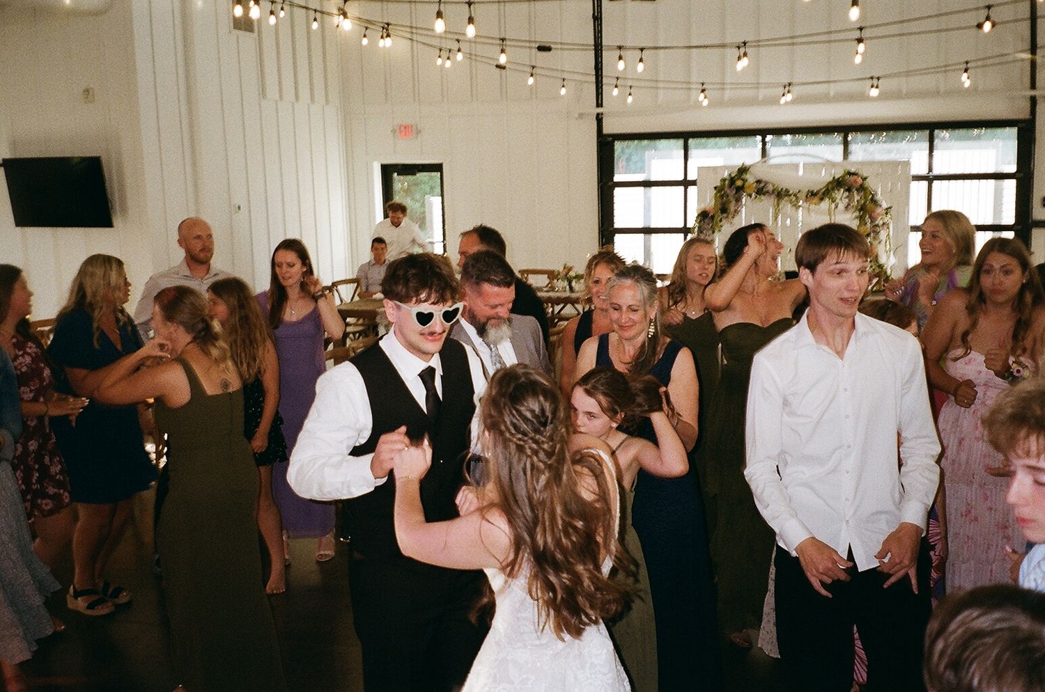 Wedding reception guests dancing joyfully in a decorated venue with string lights, including bride and groom in sunglasses.