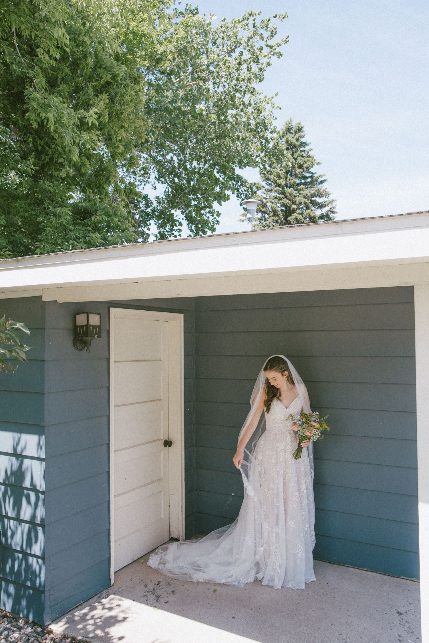 Bride in lace wedding gown and veil holding bouquet standing by gray building door with trees in background.
