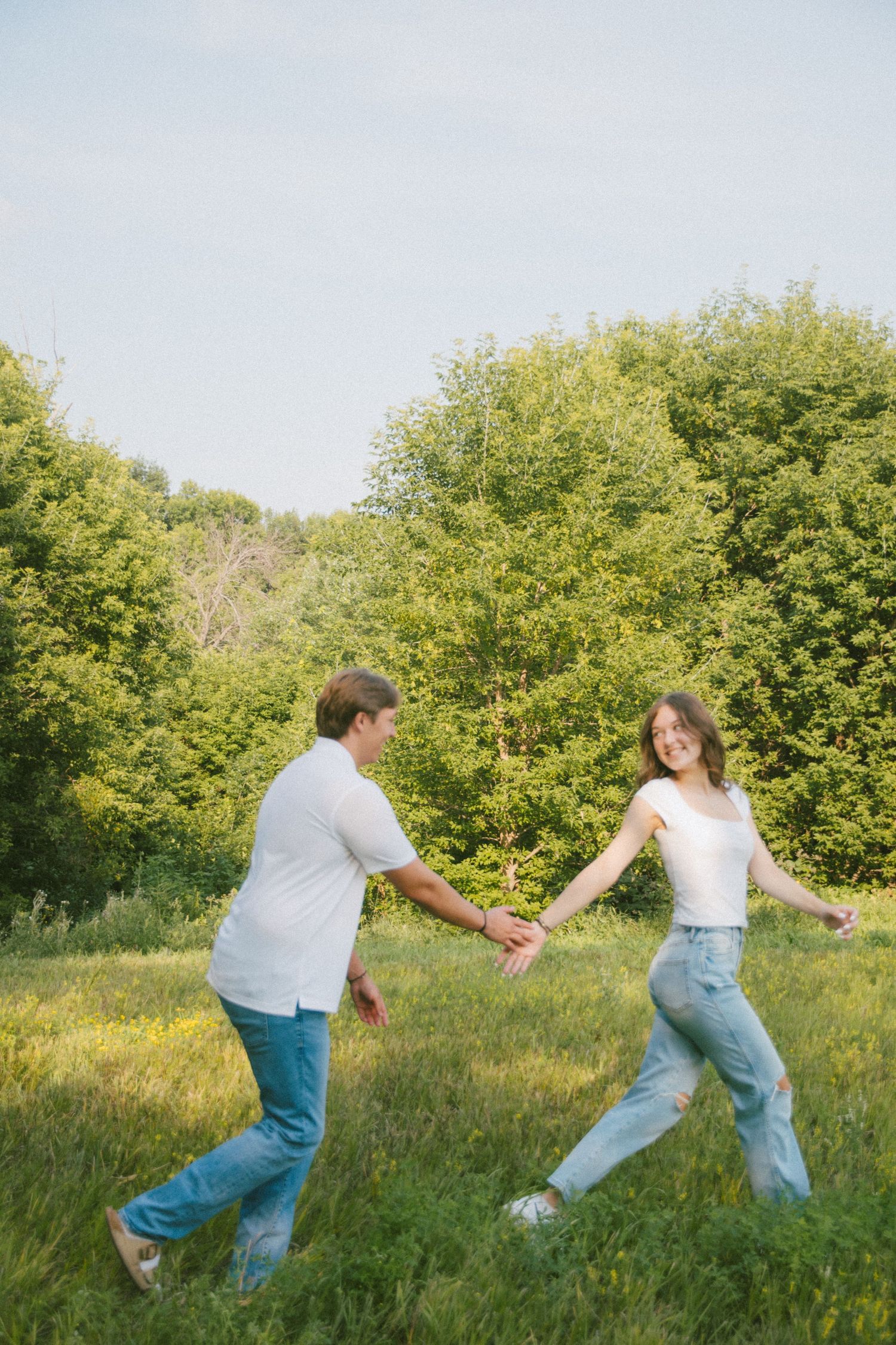 Couple holding hands and dancing playfully in a bright sunny green meadow with trees.