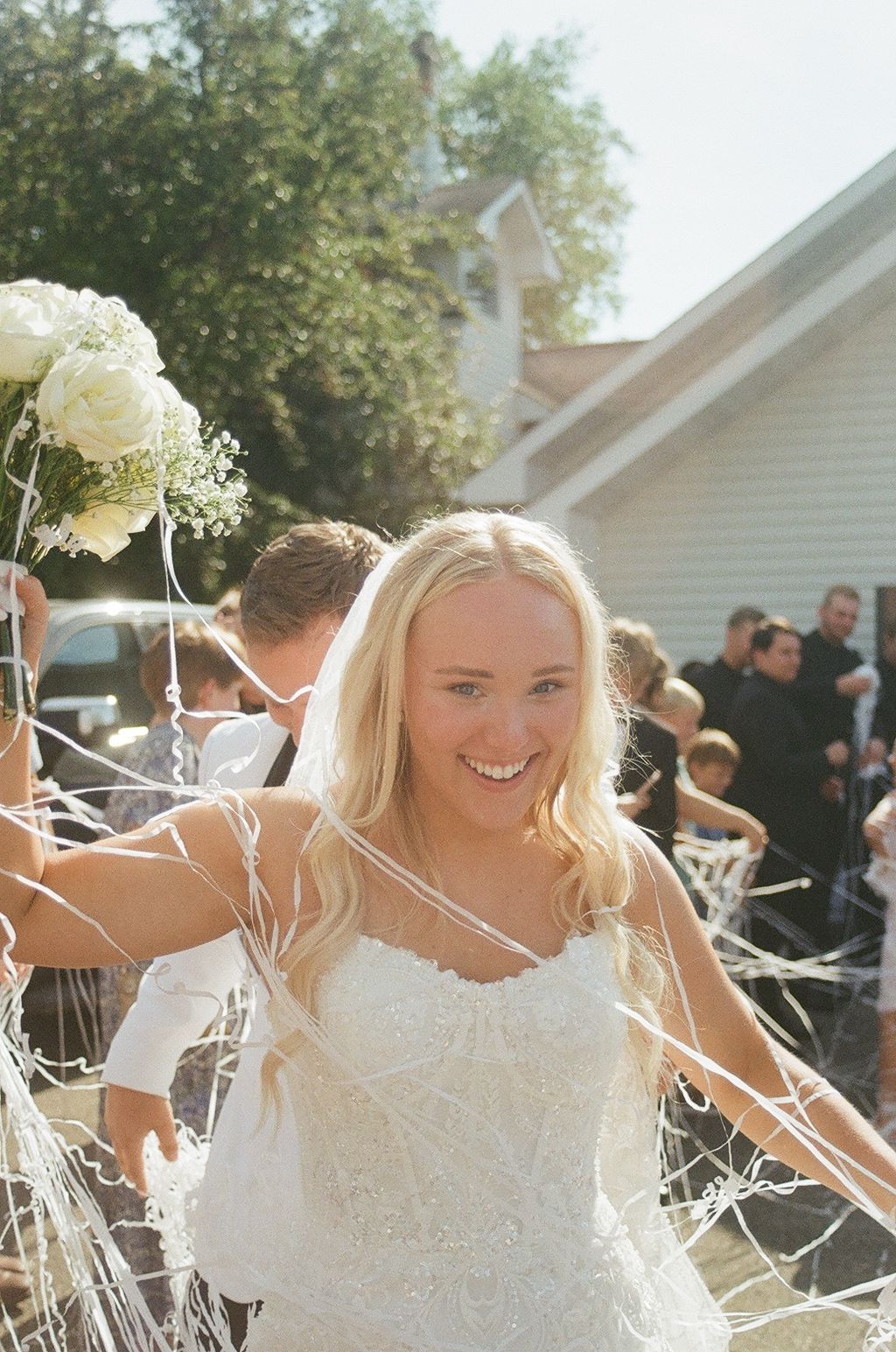 Smiling bride in white lace gown holding bouquet, surrounded by streamers during outdoor wedding celebration.