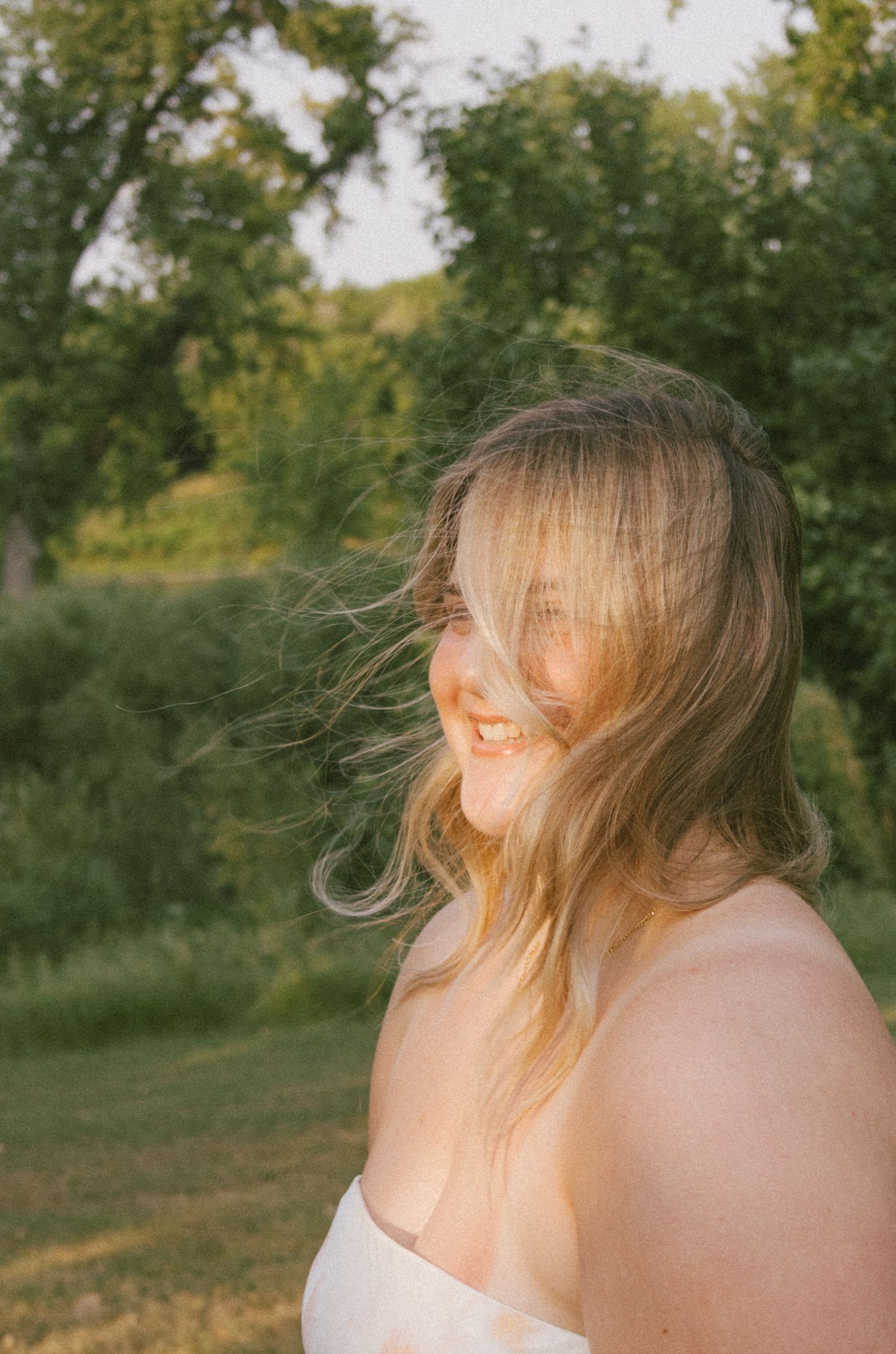 A smiling young woman with windswept blonde hair stands outdoors in golden light with lush green trees behind her.