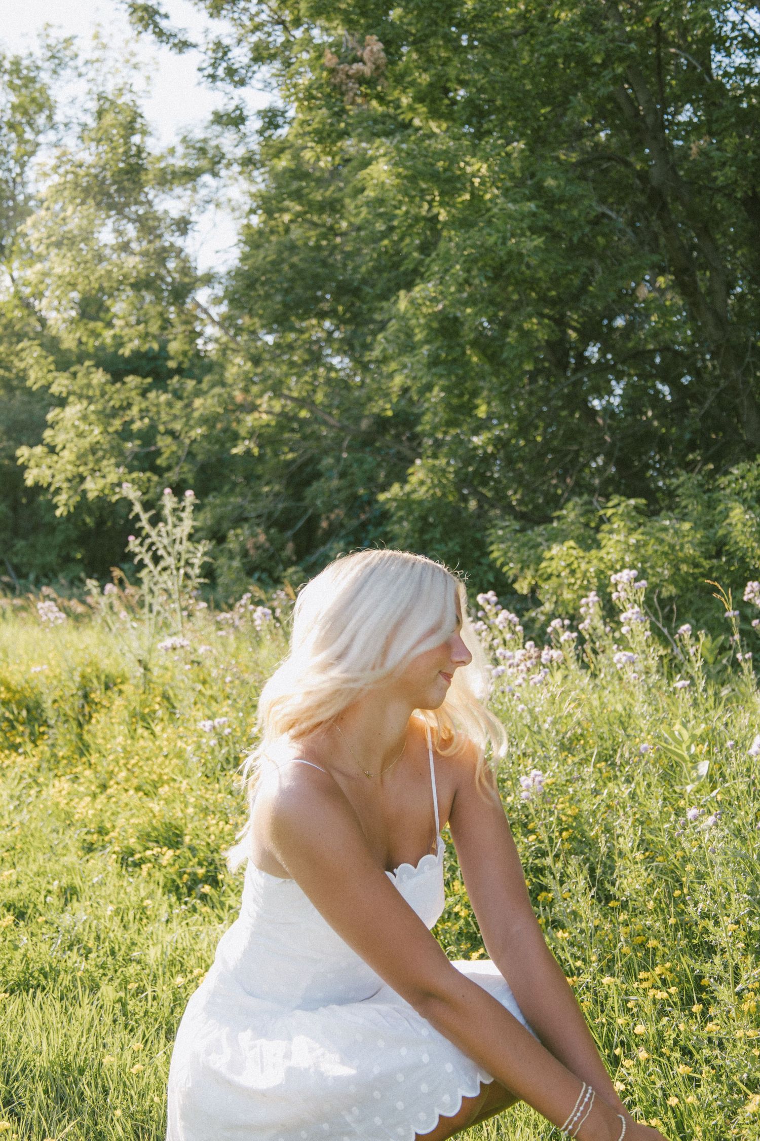 Blonde woman in white lace dress sitting in a sunny wildflower meadow surrounded by lush green trees.