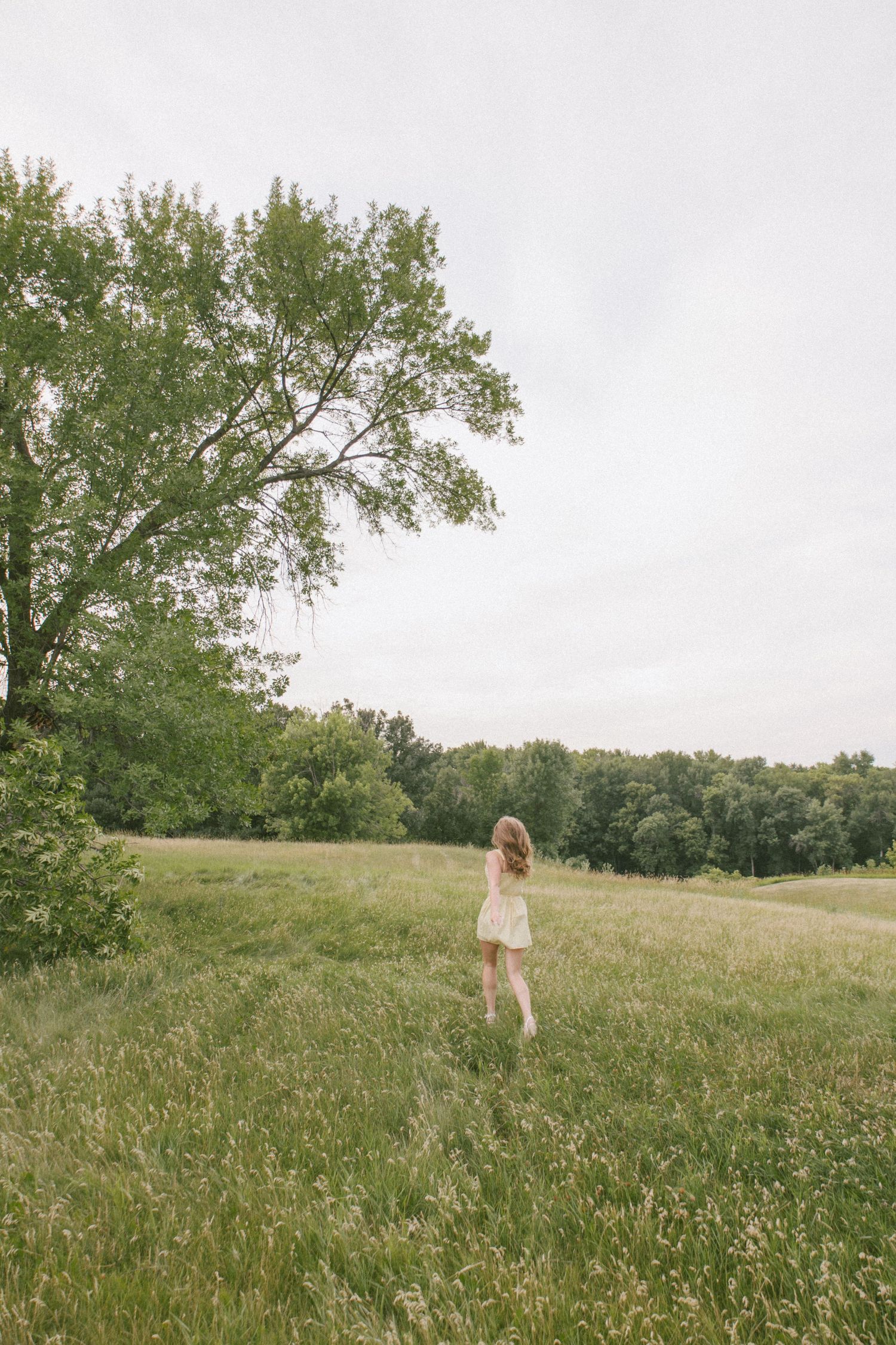 A young woman in a light dress walks through a lush green meadow with tall trees under an overcast sky.