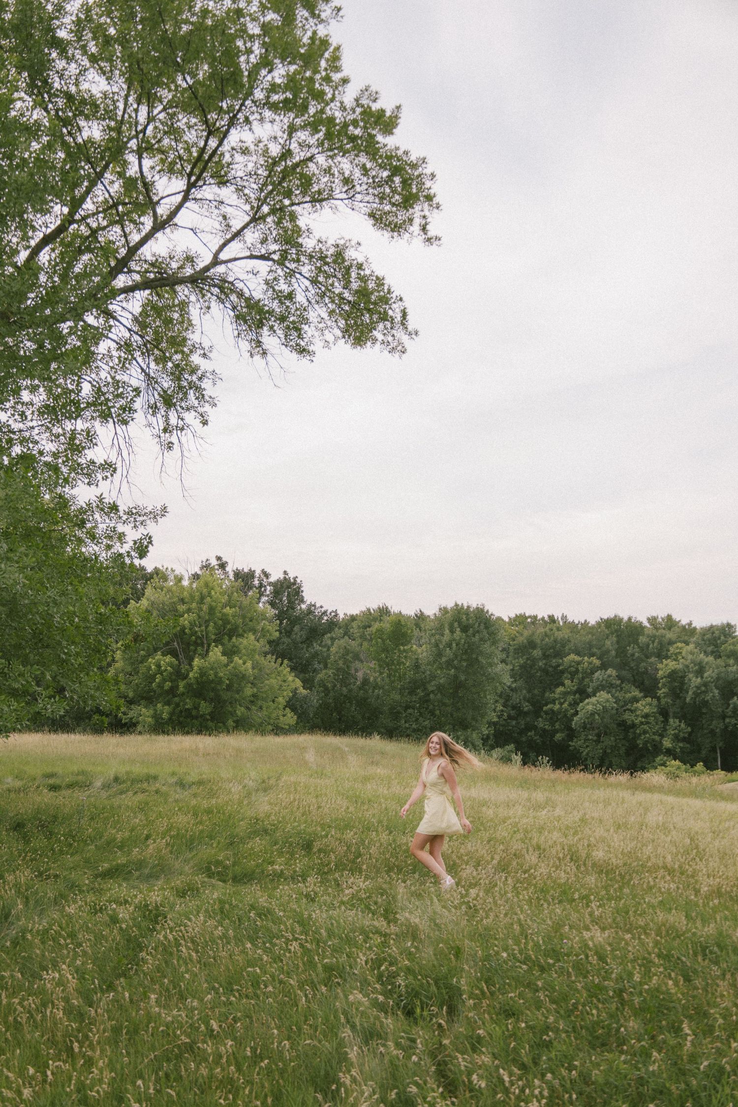 A young woman in a light dress walks through a lush green meadow surrounded by tall trees on a cloudy day.