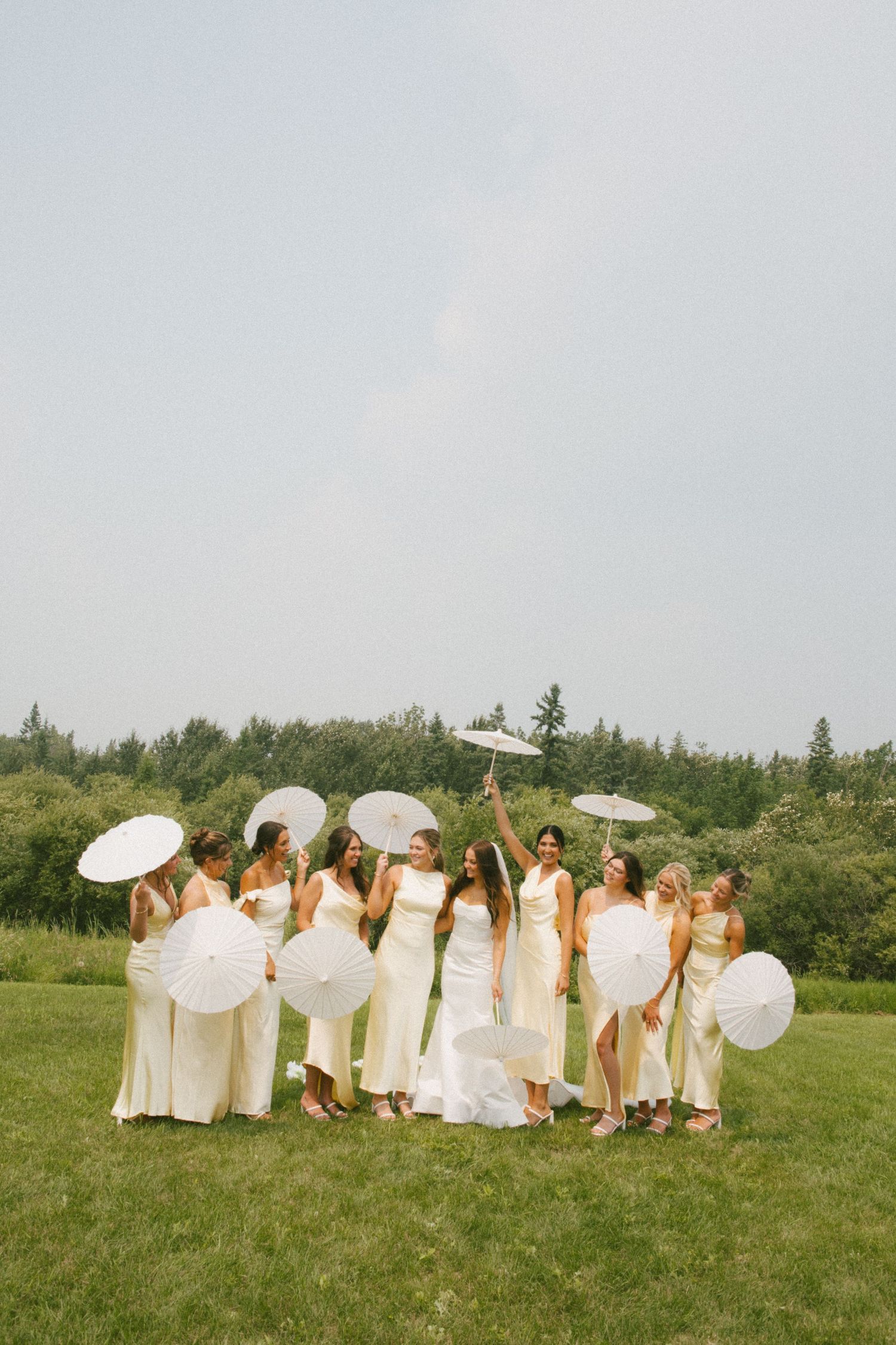 Bridal party in champagne dresses celebrating with white parasols in an open green field.