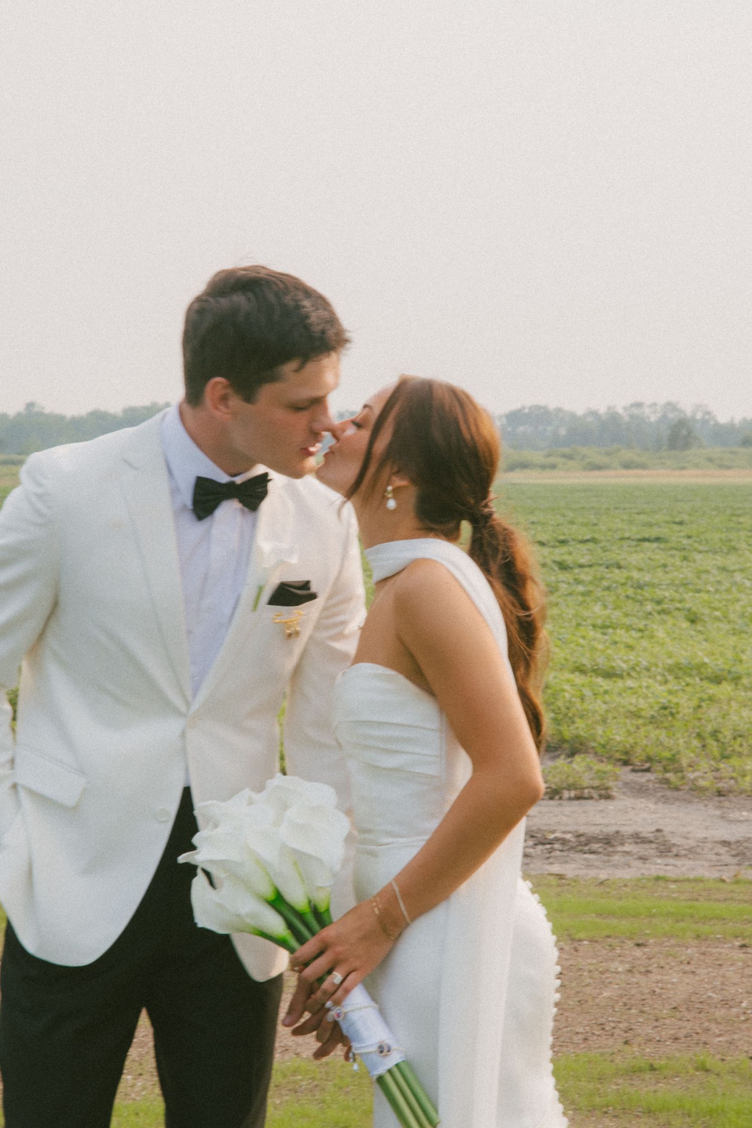 A bride and groom share a kiss in front of a green field, she holds white calla lilies.