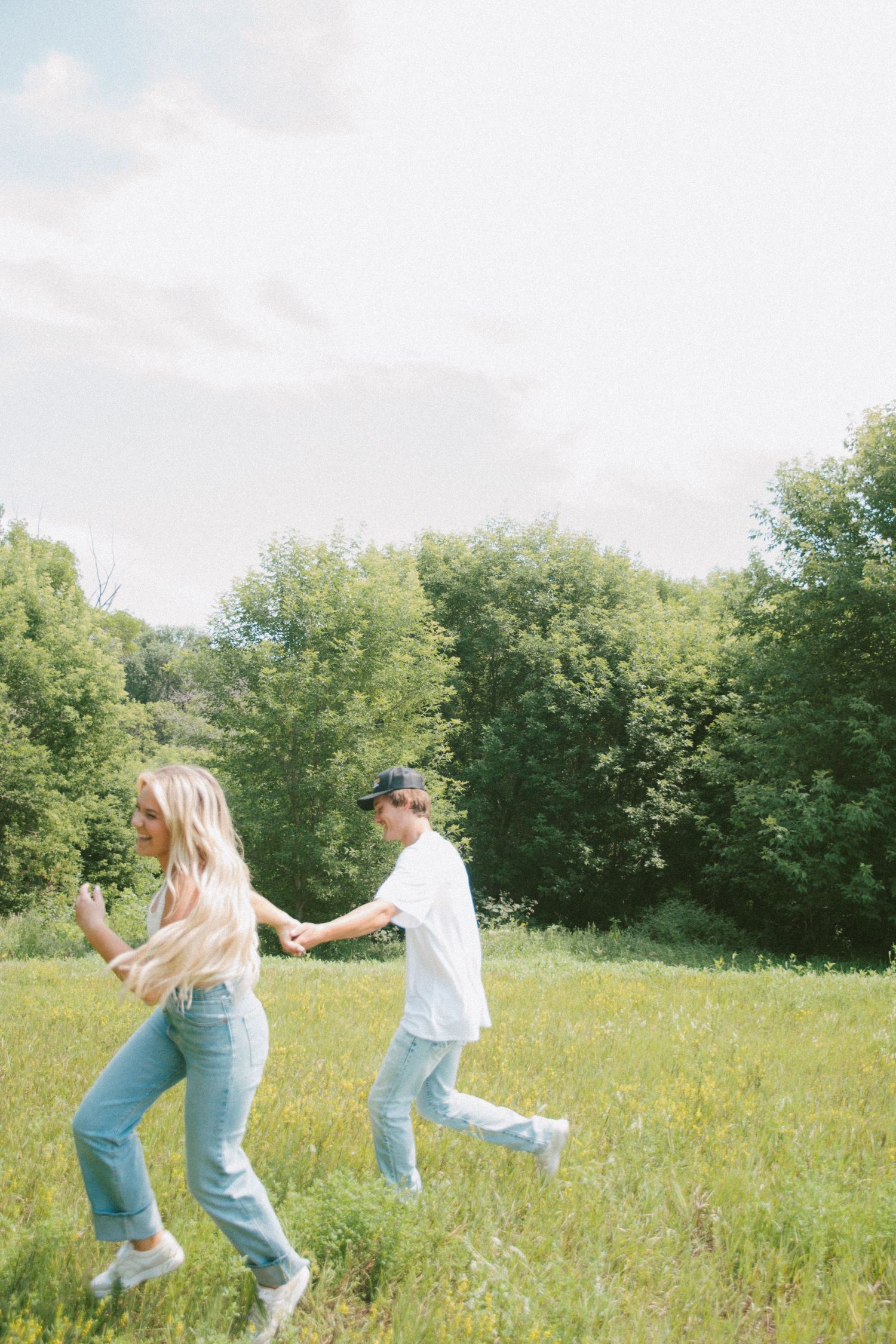Couple running playfully through a sunny green meadow holding hands on a cloudy day.