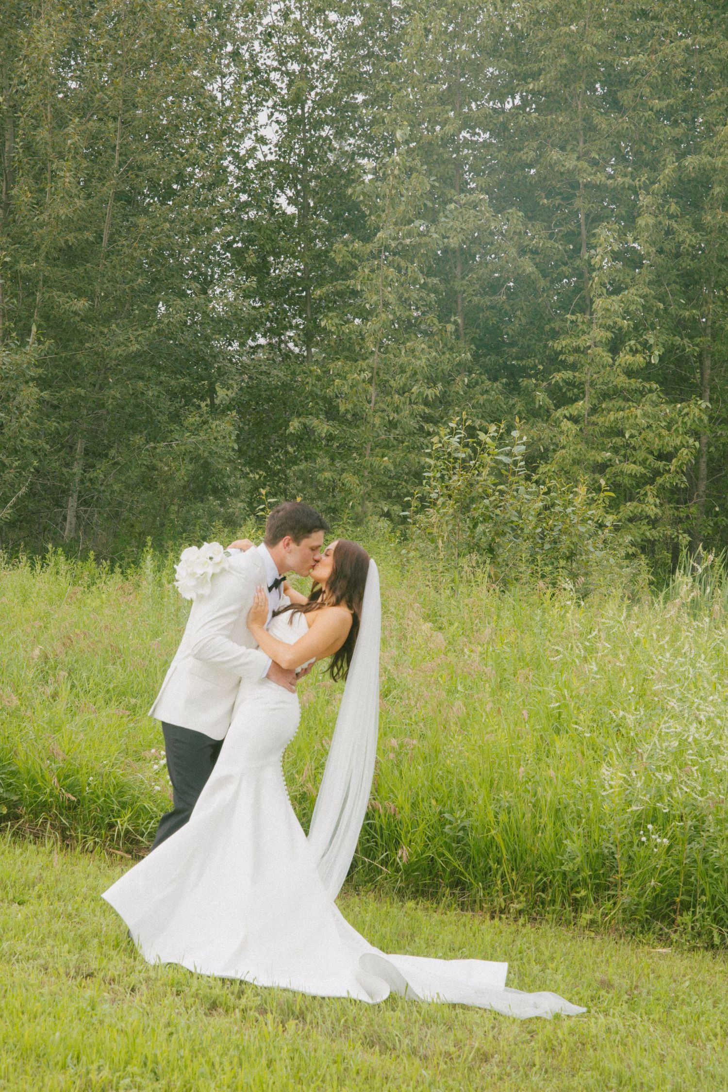 Bride and groom sharing a passionate kiss in a lush green meadow, brides long veil trailing behind.