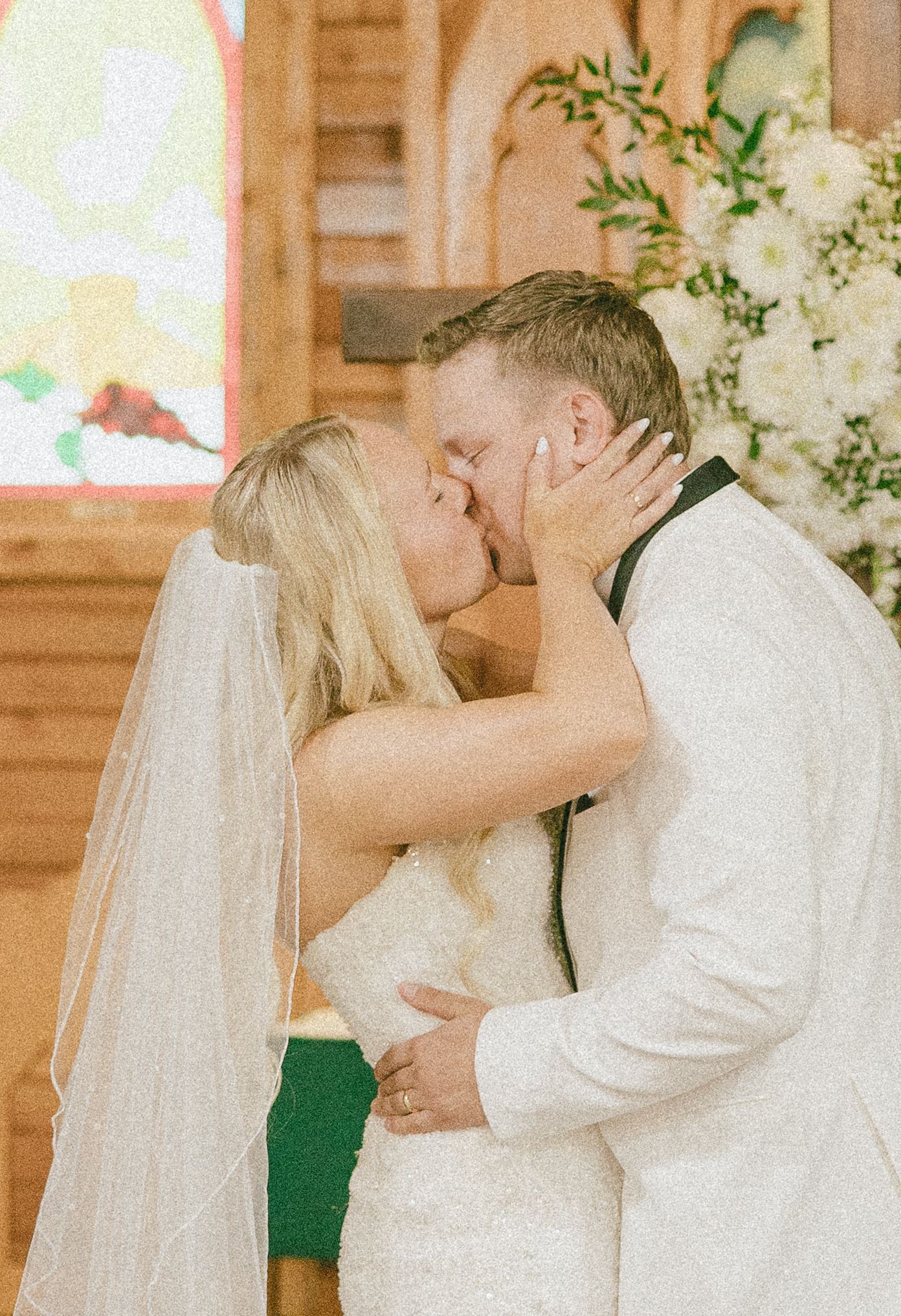 Bride and groom share first kiss at church altar, bride in white gown with veil, groom in white suit.