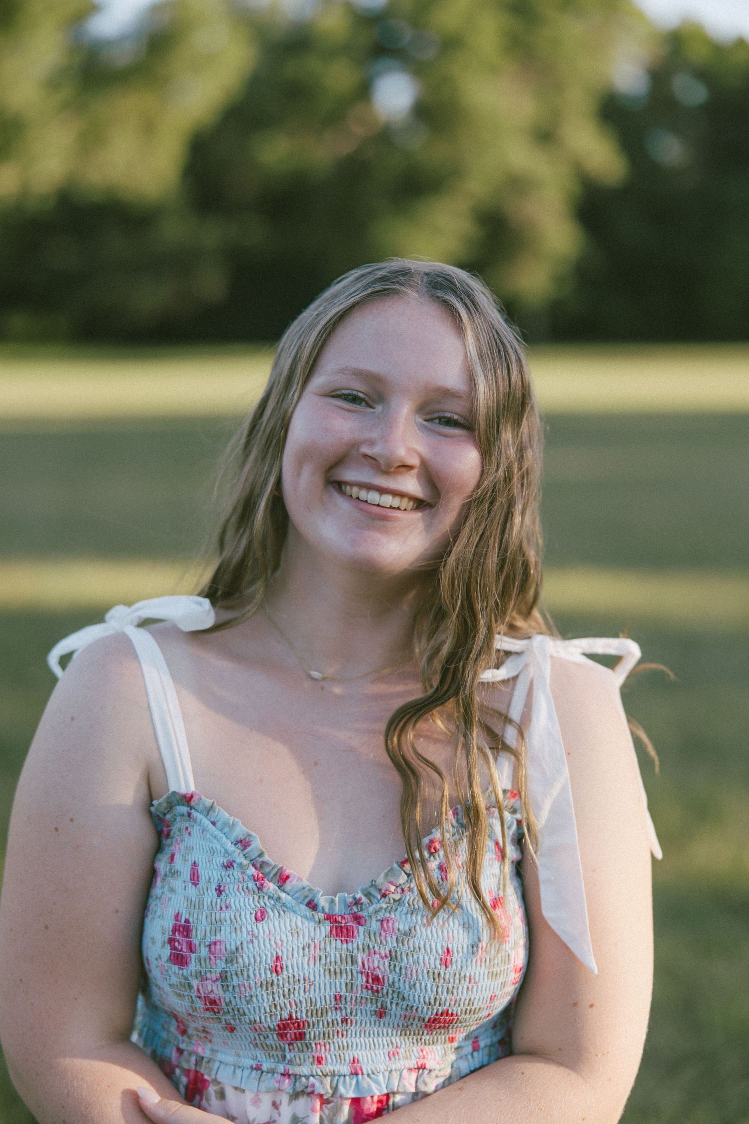 Smiling young woman in floral sundress with bow straps standing outdoors in a sunny green field.