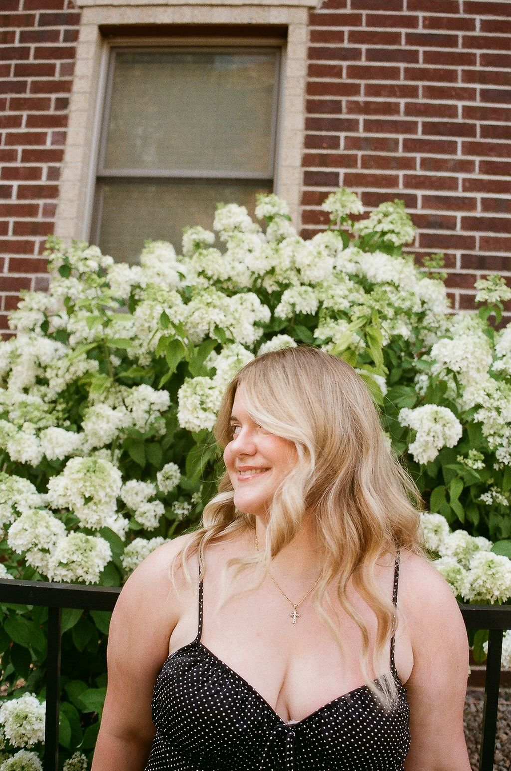 Smiling blonde woman in black polka dot dress standing in front of white hydrangea bushes and brick building.