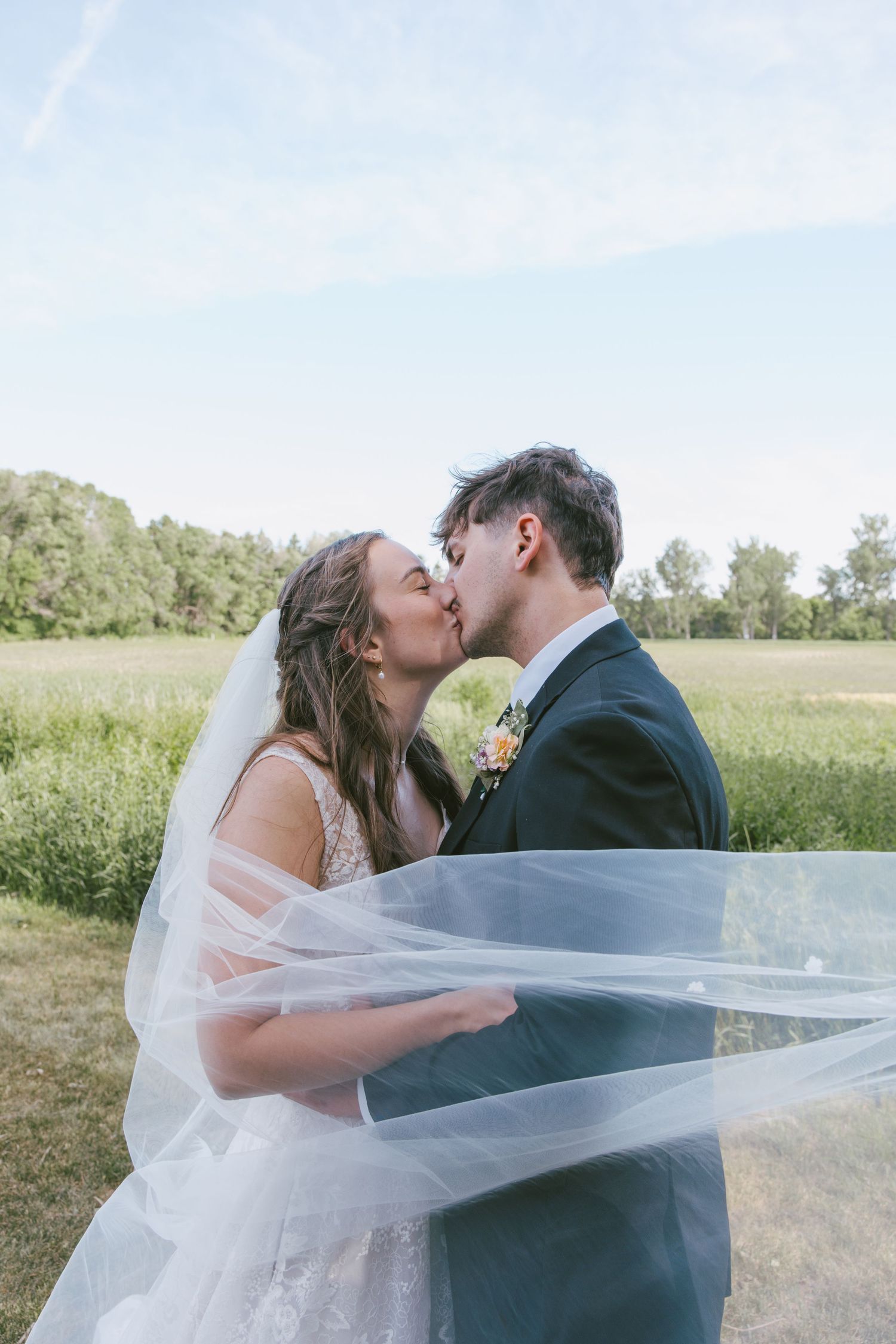 Bride and groom share a romantic kiss outdoors, with her veil flowing in the wind against a lush green field.