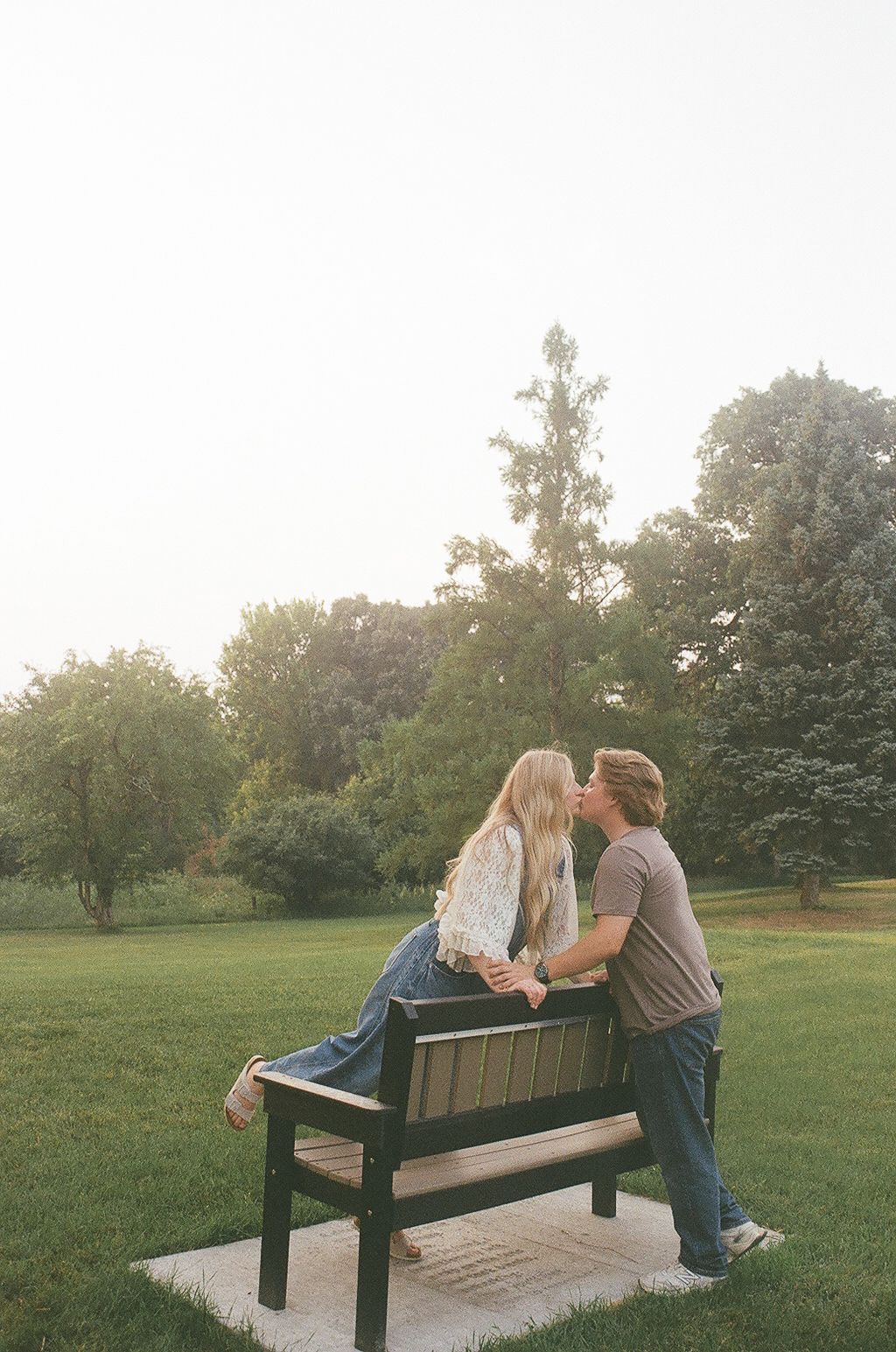A couple shares a romantic kiss on a park bench surrounded by lush green trees and grass at sunset.
