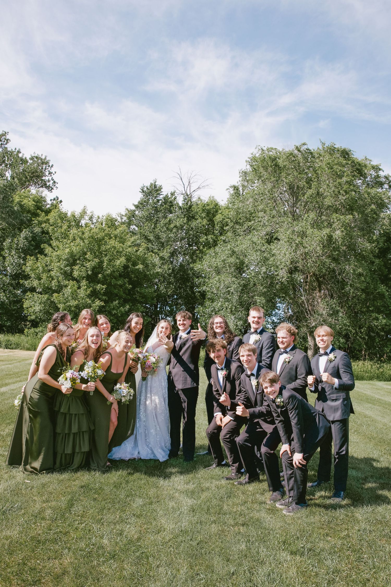 Wedding party in olive green and black tuxedos poses joyfully on green lawn with lush trees in background.