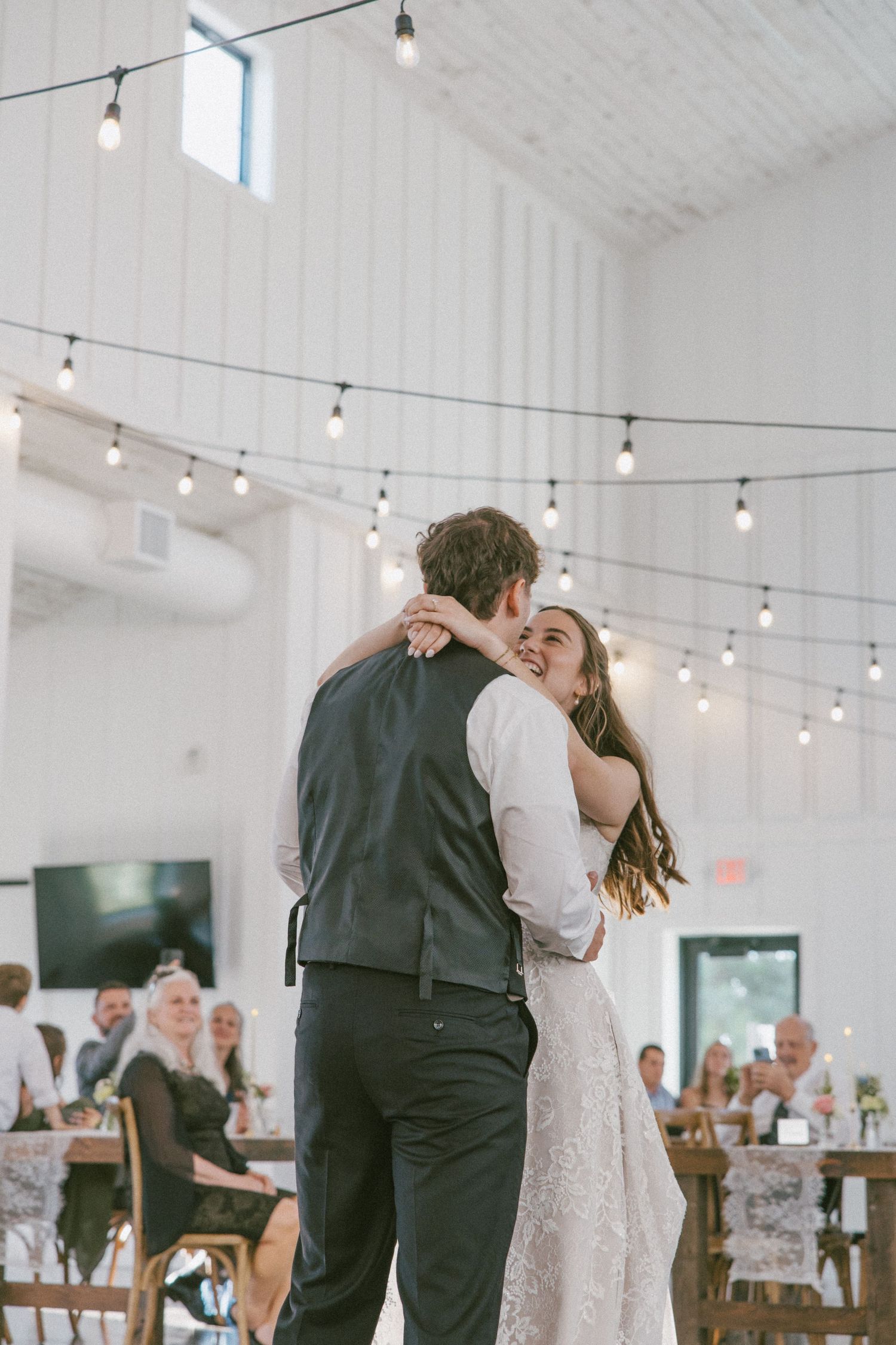 Bride and groom share their first dance under string lights in a white barn wedding venue with guests seated in background.