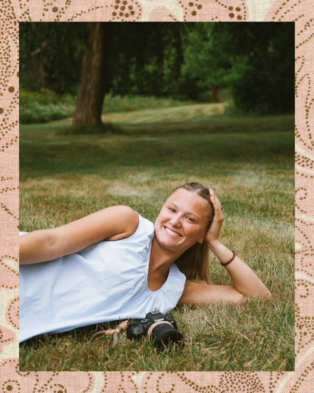 Young woman smiling while lying on grass in a park, holding a camera, wearing a light blue sleeveless dress.
