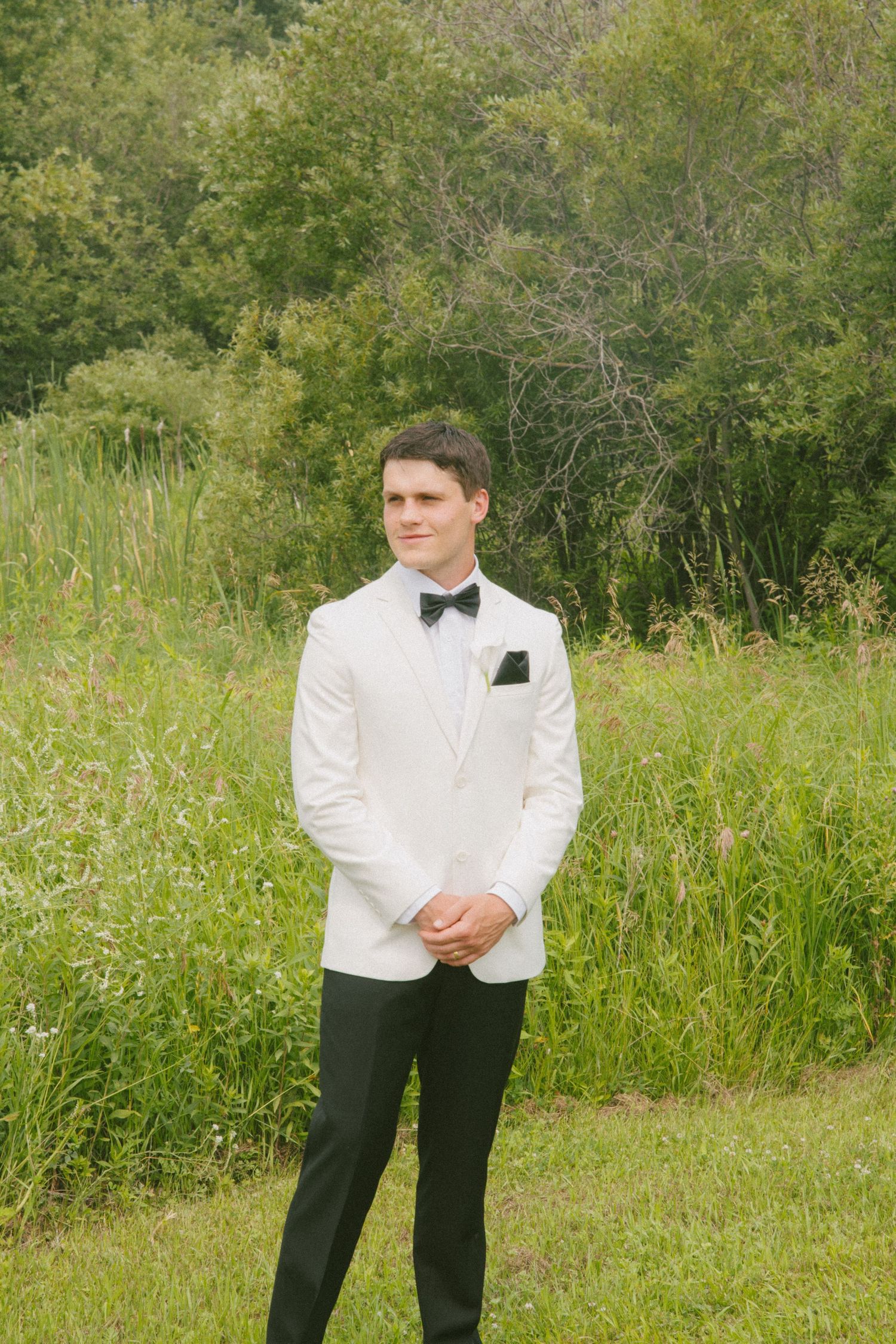 Handsome groom in white tuxedo jacket and black bow tie standing in front of lush green foliage.
