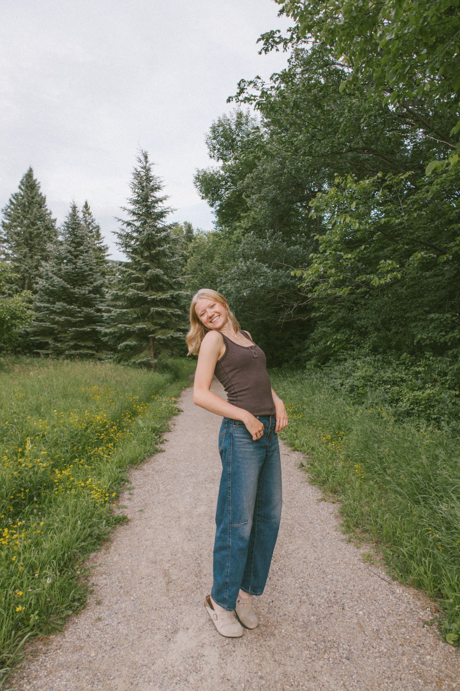 Smiling blonde woman in brown tank top and jeans standing on a gravel path surrounded by lush green trees.