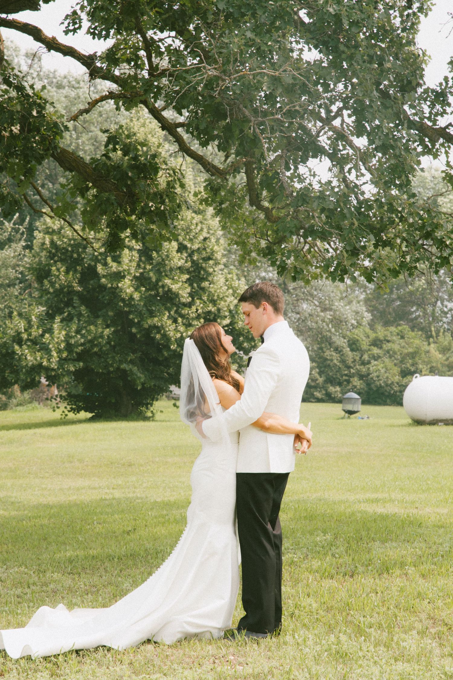 Bride in white gown with veil and groom in white tuxedo sharing a romantic moment under a large tree.