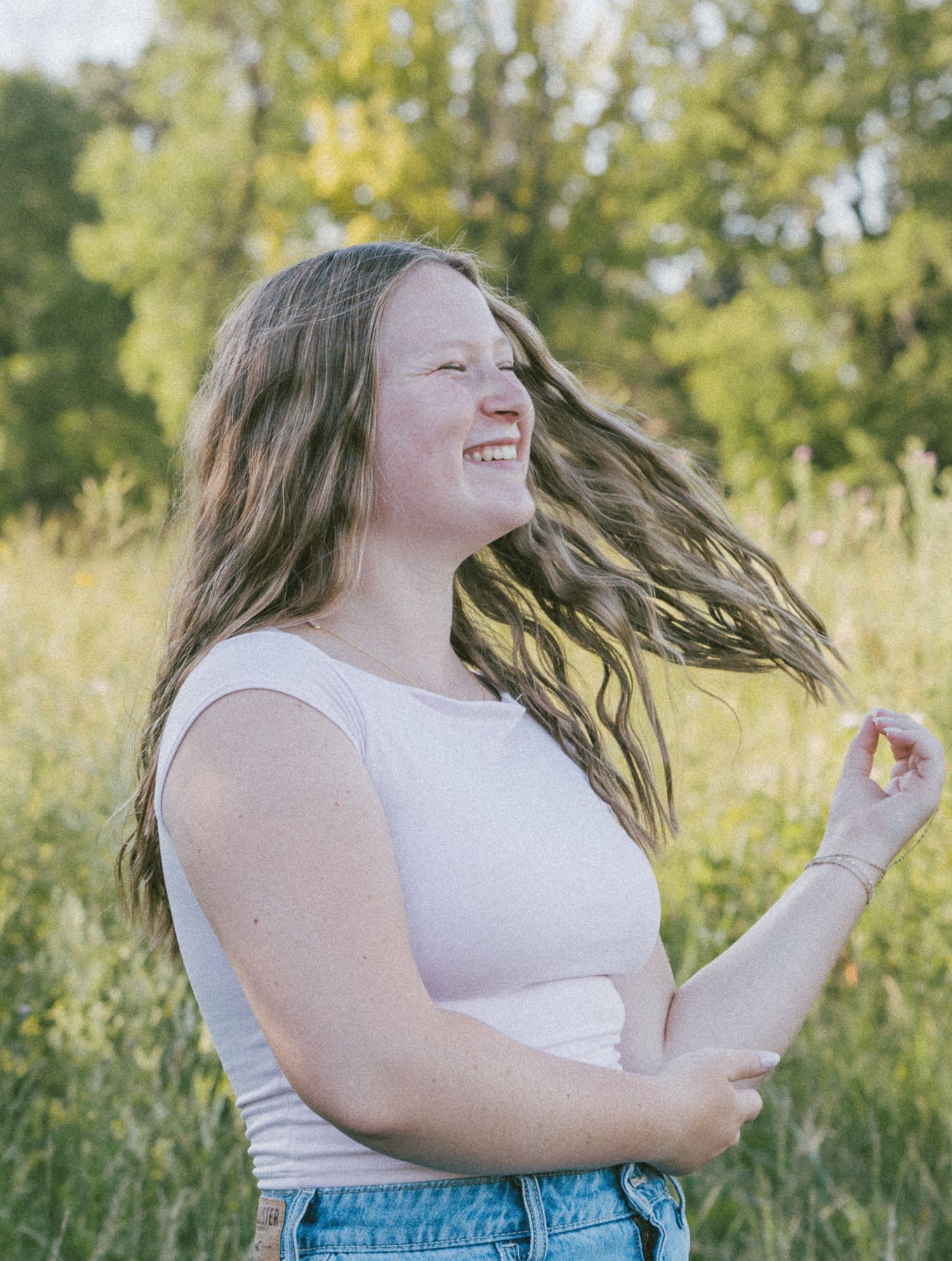 Young woman with long hair laughing joyfully outdoors in a sunny green meadow, wearing a white top and denim shorts.