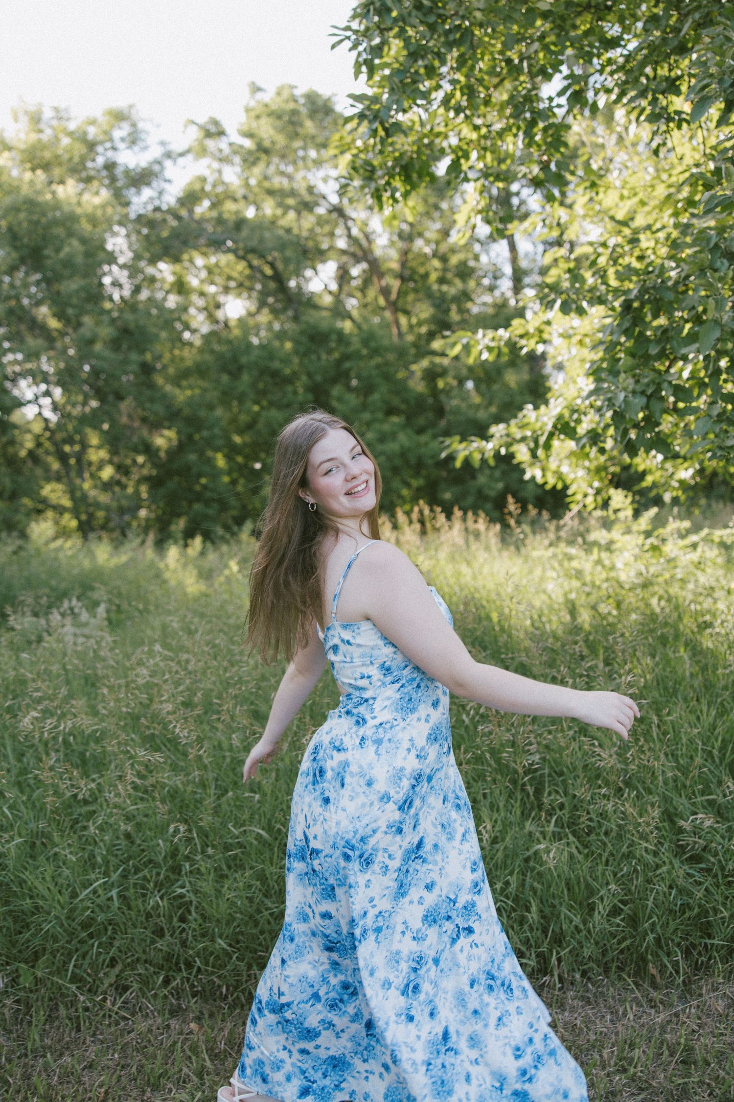 Young woman in blue floral maxi dress twirling in a lush green meadow, smiling over her shoulder on a sunny day.