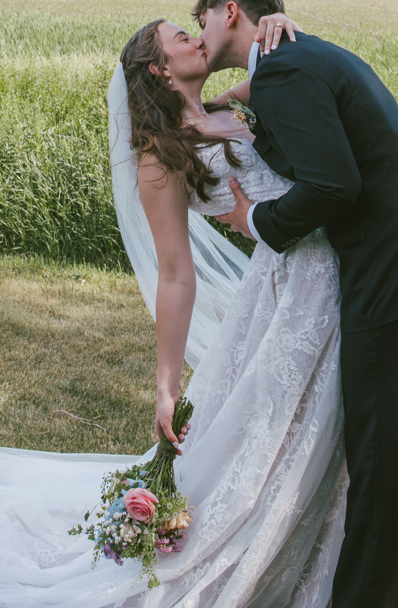 Bride and groom sharing a romantic kiss outdoors, bride holding a colorful wildflower bouquet in a lace wedding gown.
