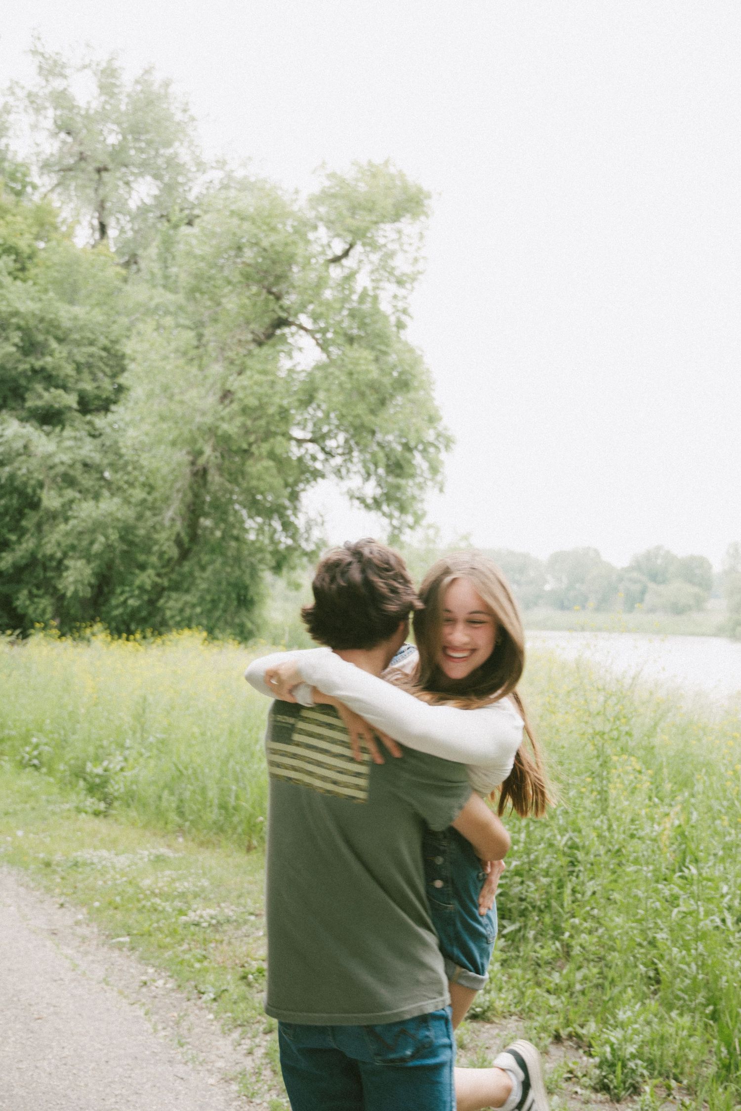 Young couple embracing and smiling on a scenic path beside wildflowers and a river.