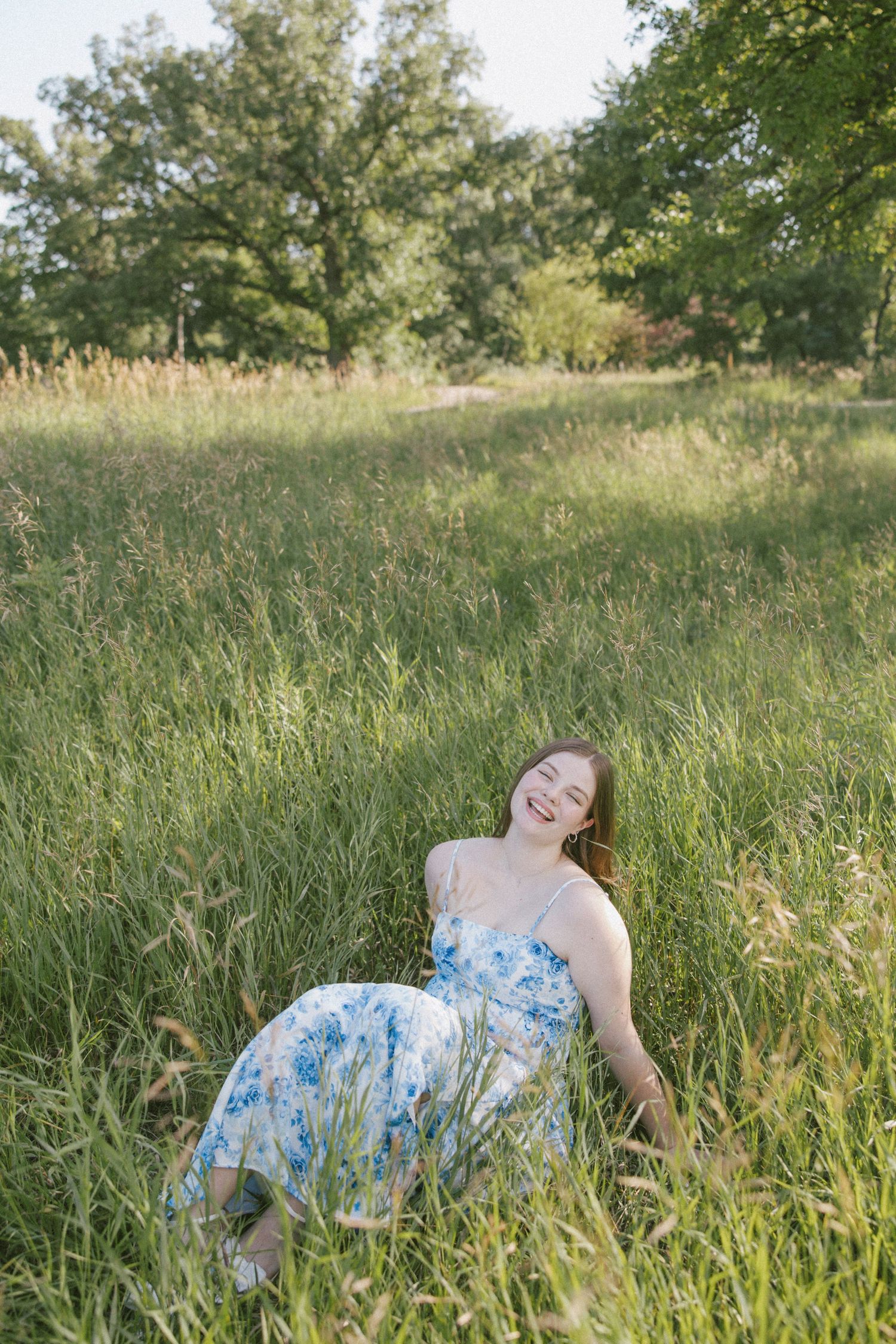 Young woman in blue floral sundress laughing joyfully while sitting in tall green grass field with trees in background.