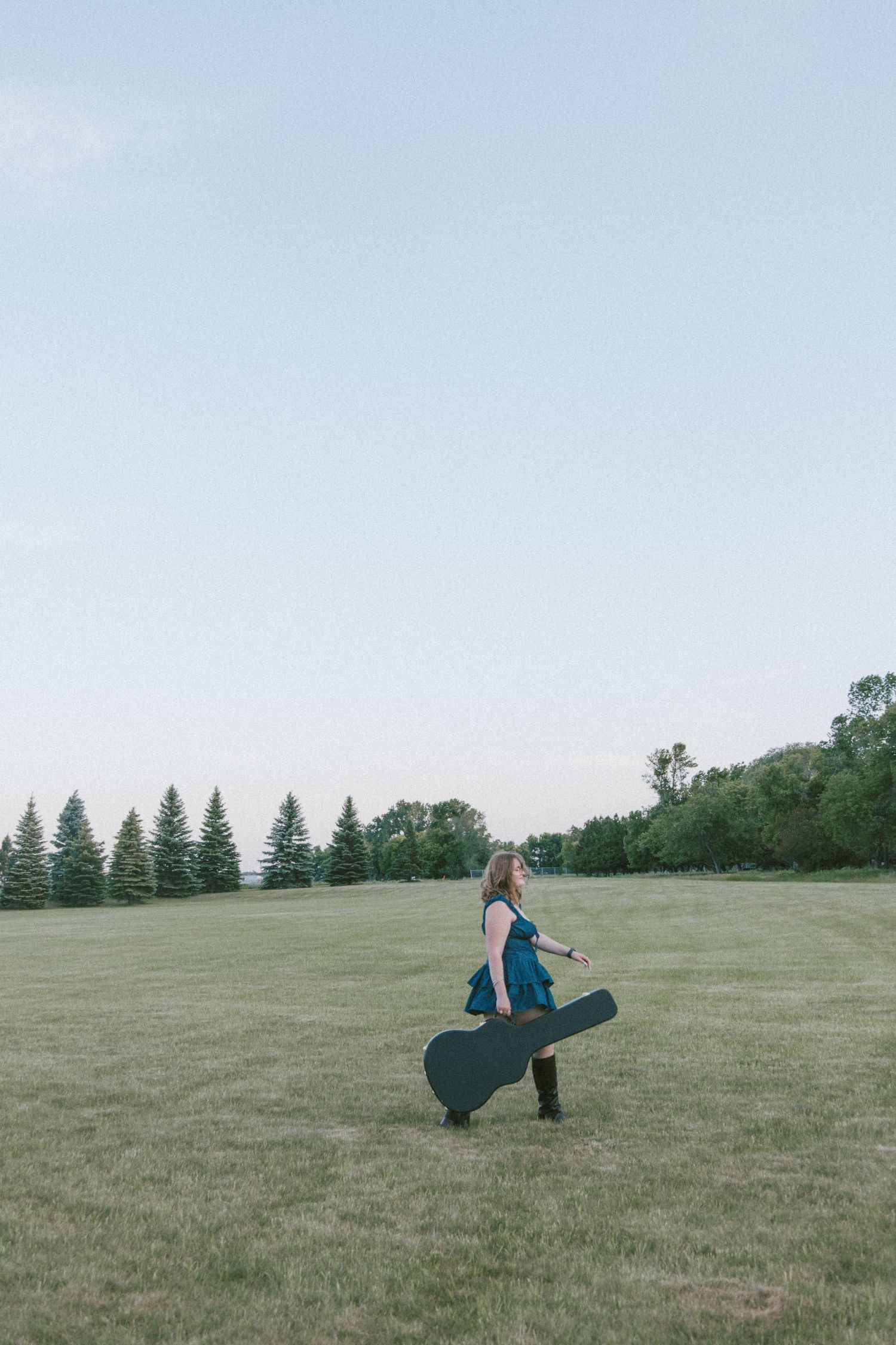 Woman in teal dress carrying guitar case walks across green field with evergreen trees and clear sky.