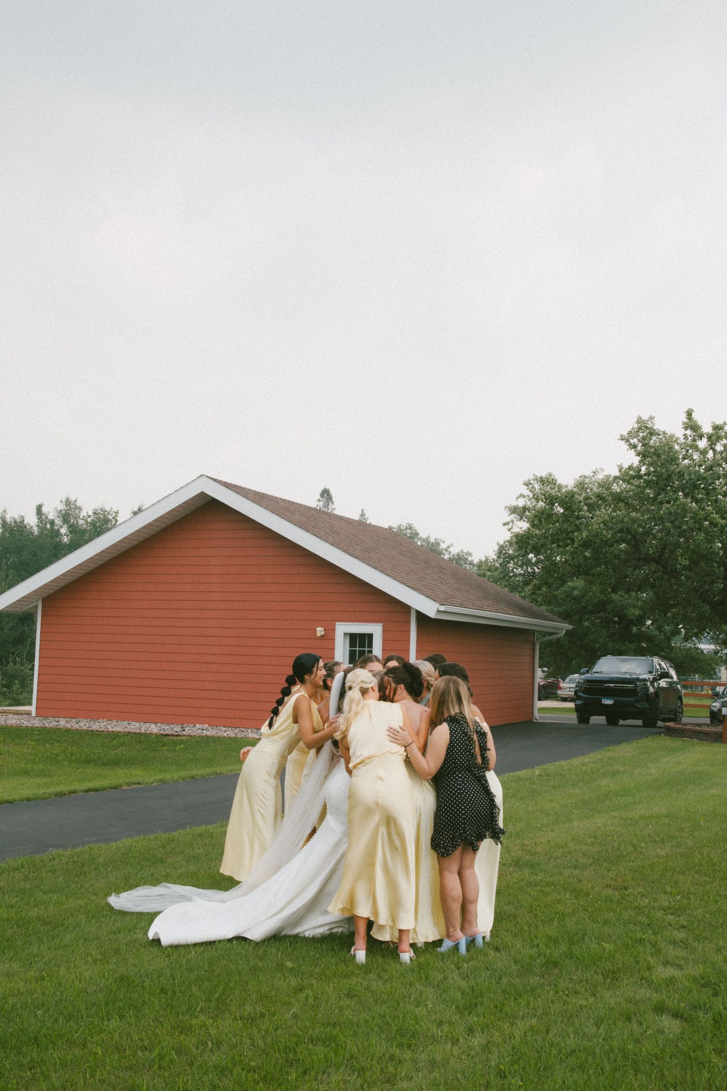 Bride and bridesmaids in champagne gowns sharing a group hug outside a red barn on wedding day.