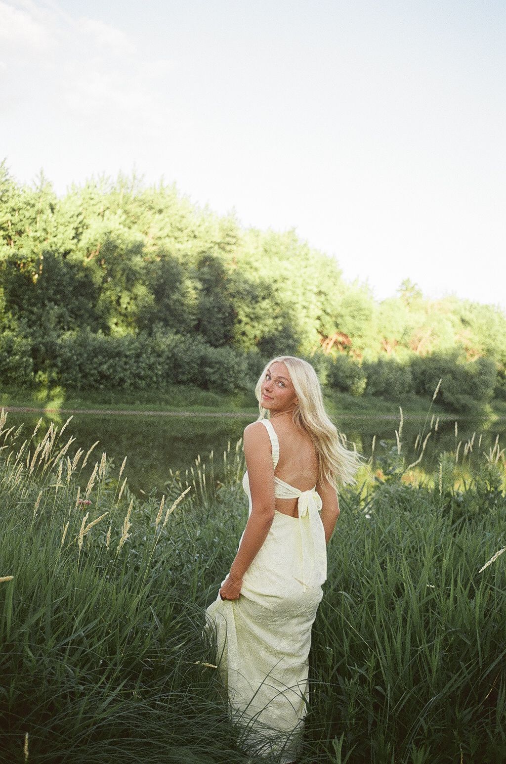 Blonde woman in white backless dress standing in lush green field near water surrounded by tall grass and trees.