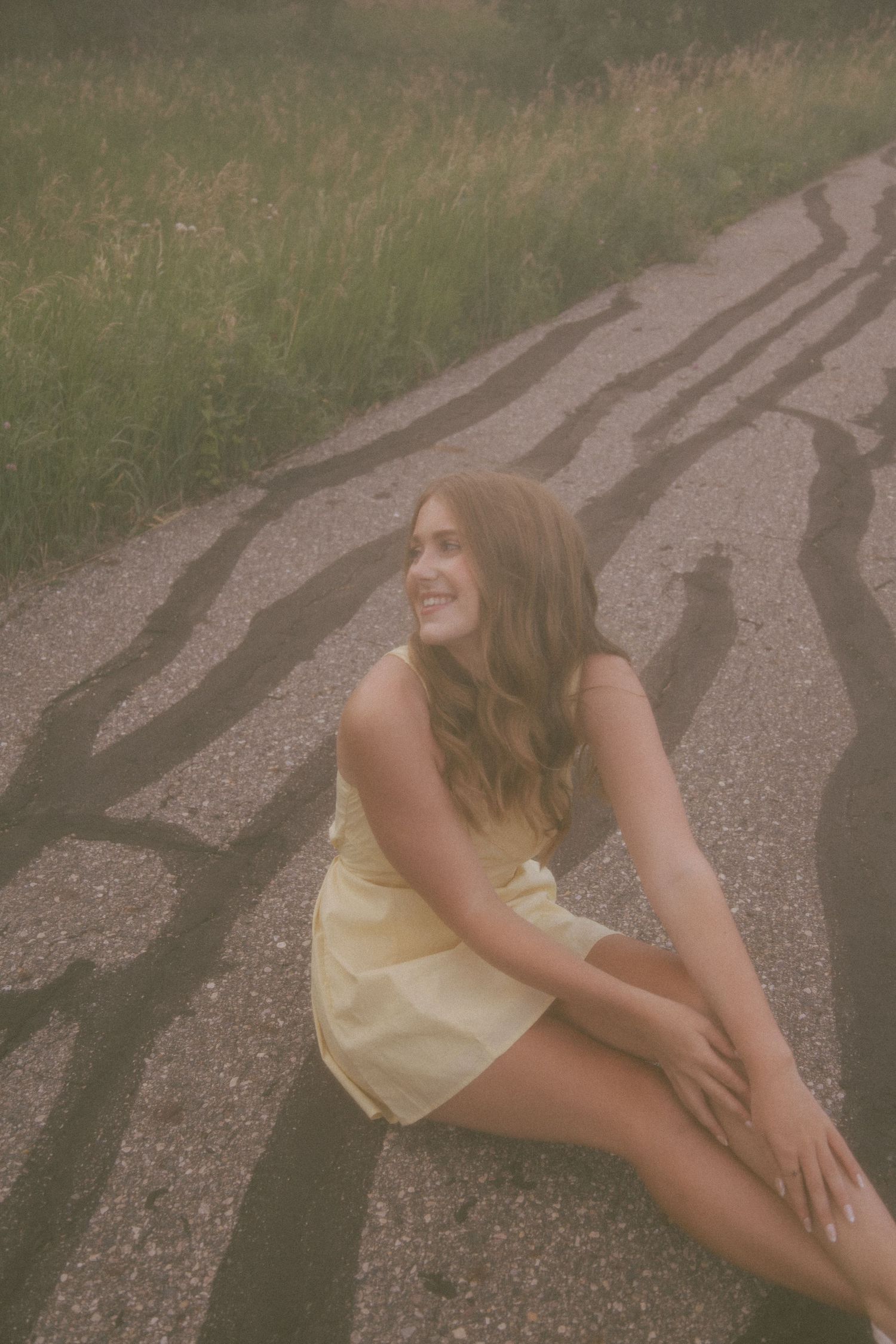 Young woman in yellow dress sitting on a sunlit road surrounded by grass and long shadows.