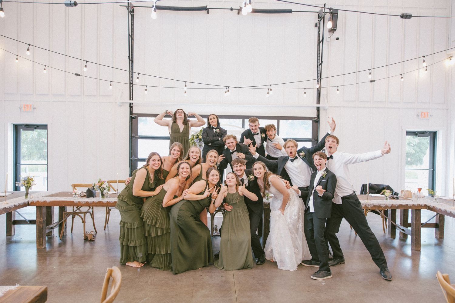 Wedding party posing joyfully in a white barn reception venue with string lights and wooden tables.