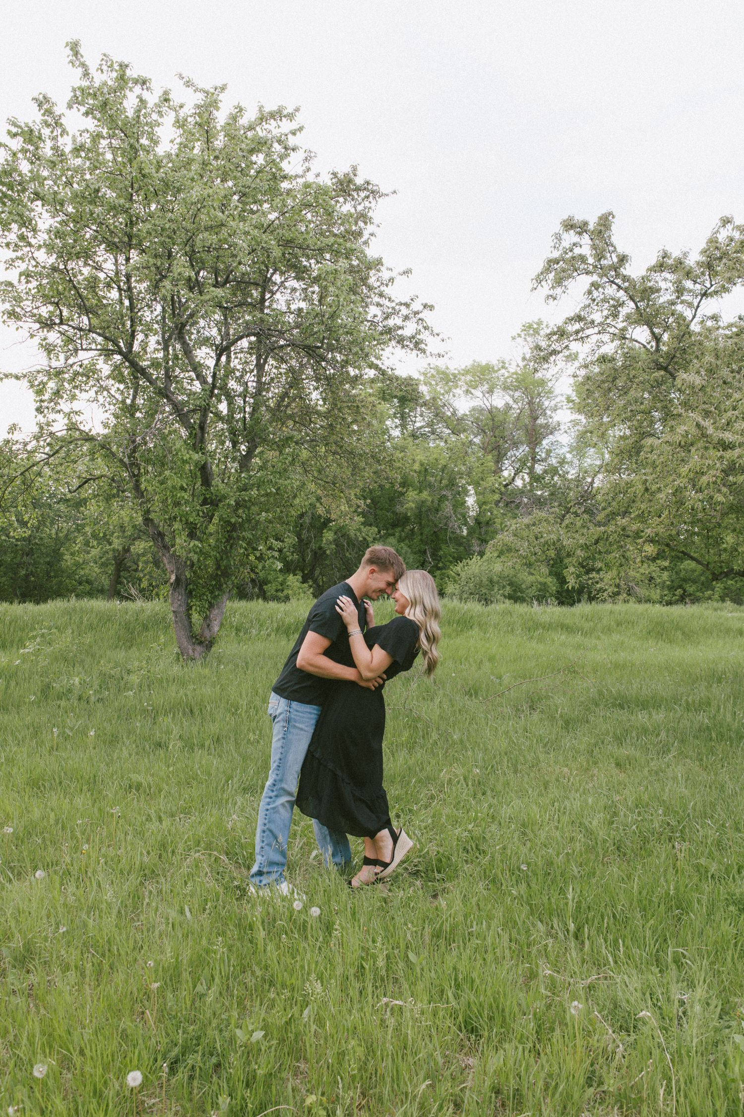Couple embracing and kissing in a lush green meadow with blooming trees, wearing black outfits and jeans.
