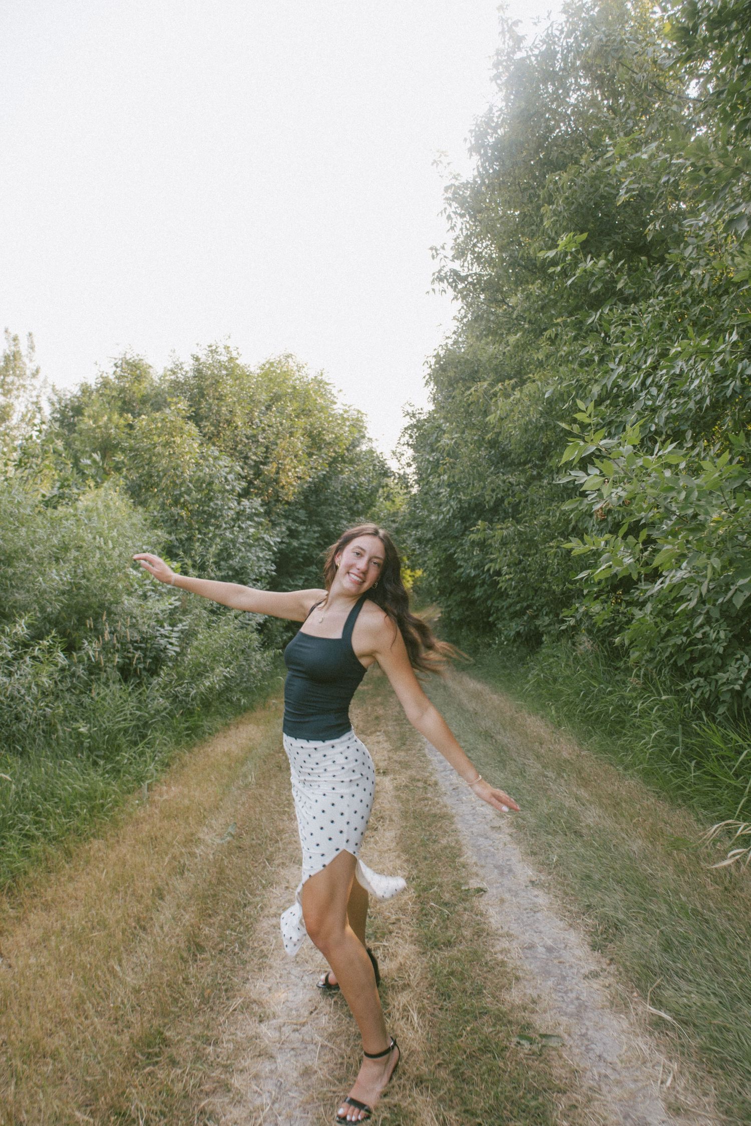 Woman in black top and polka dot skirt dancing joyfully on a rural dirt path surrounded by lush green trees.